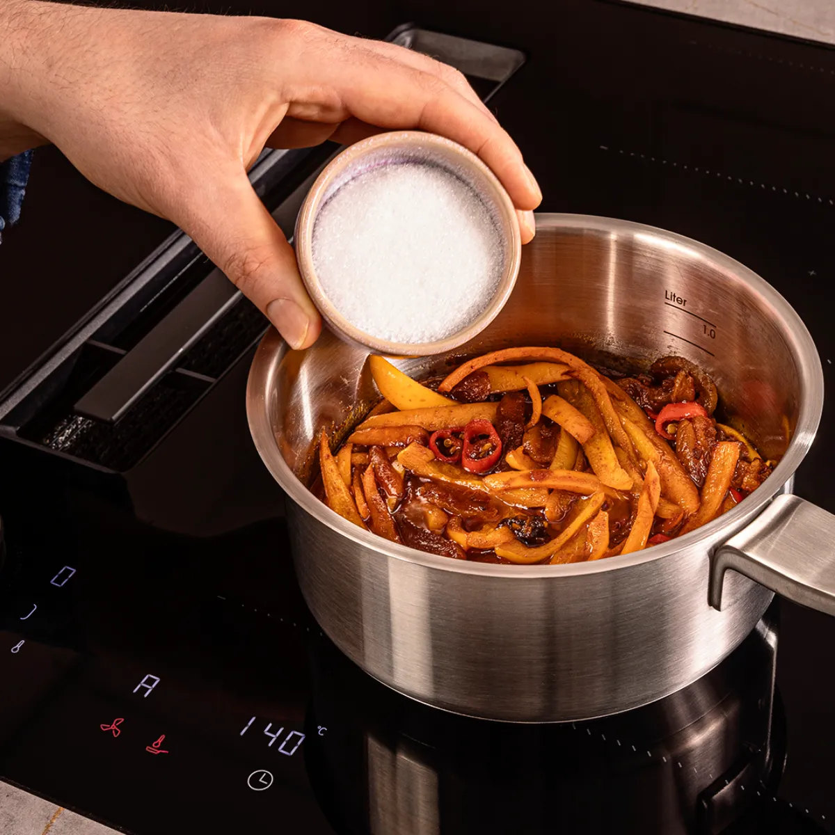 Hand adding salt to a pot with cooked sliced vegetables and chili peppers on a stovetop set to 140°C.