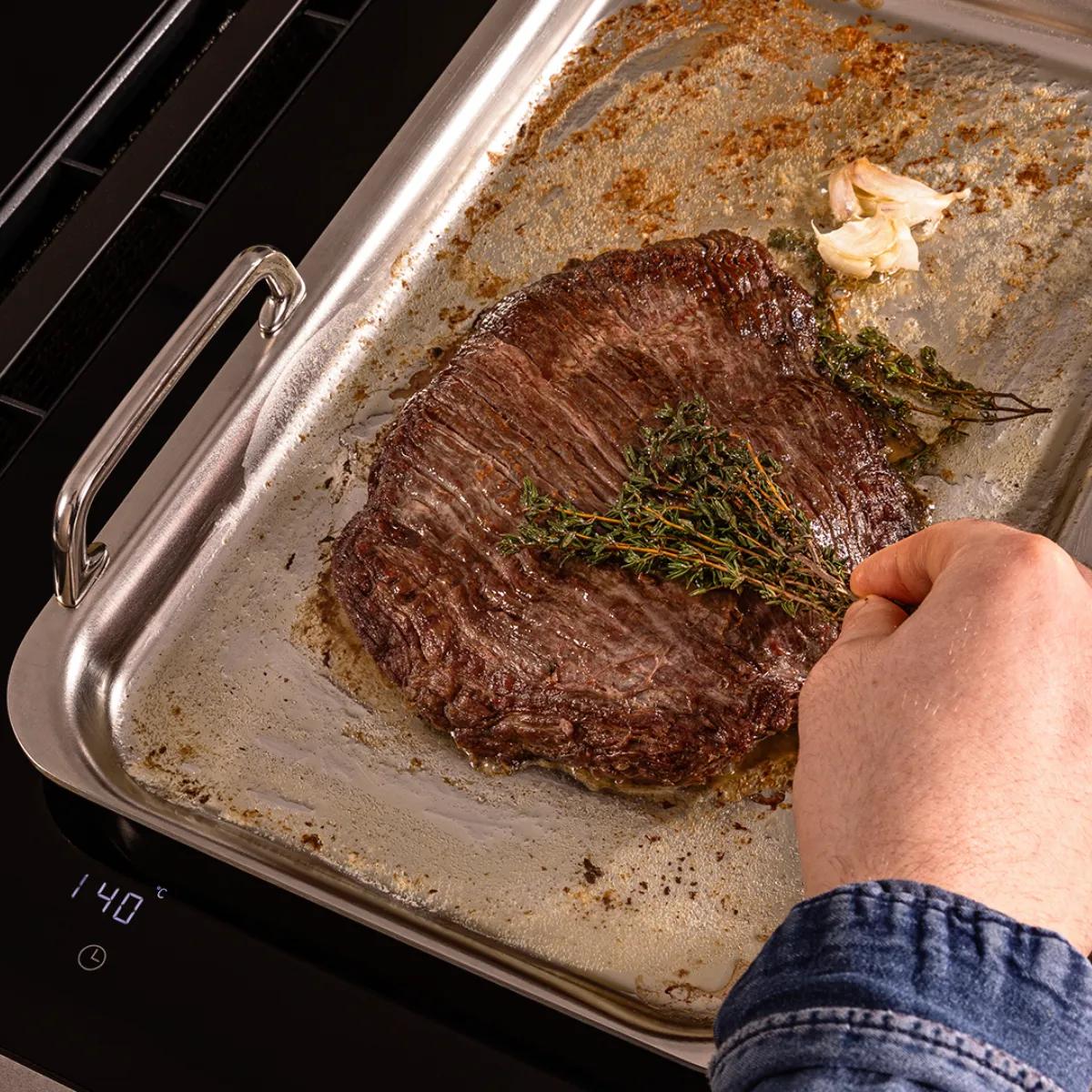 Hand cooking beef steak with fresh thyme and garlic in a metal roasting pan on an induction stove showing 140°C.