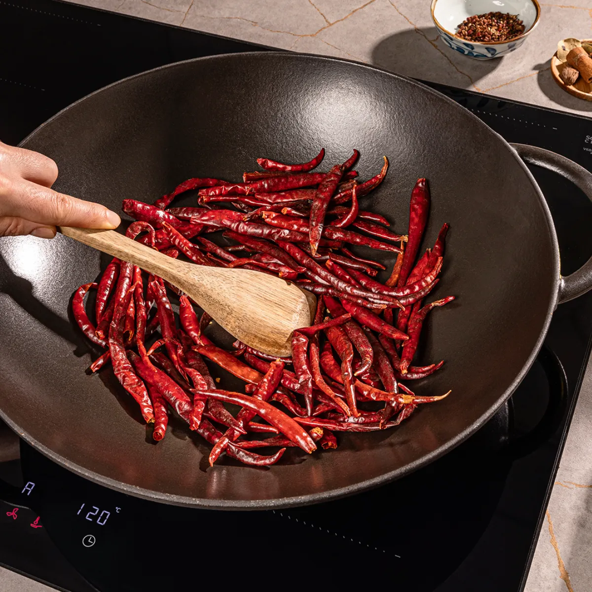 In einem schwarzen Wok auf einem Induktionsherd werden getrocknete rote Chilischoten von Hand gerührt, während daneben kleine Schälchen mit Gewürzen stehen.
