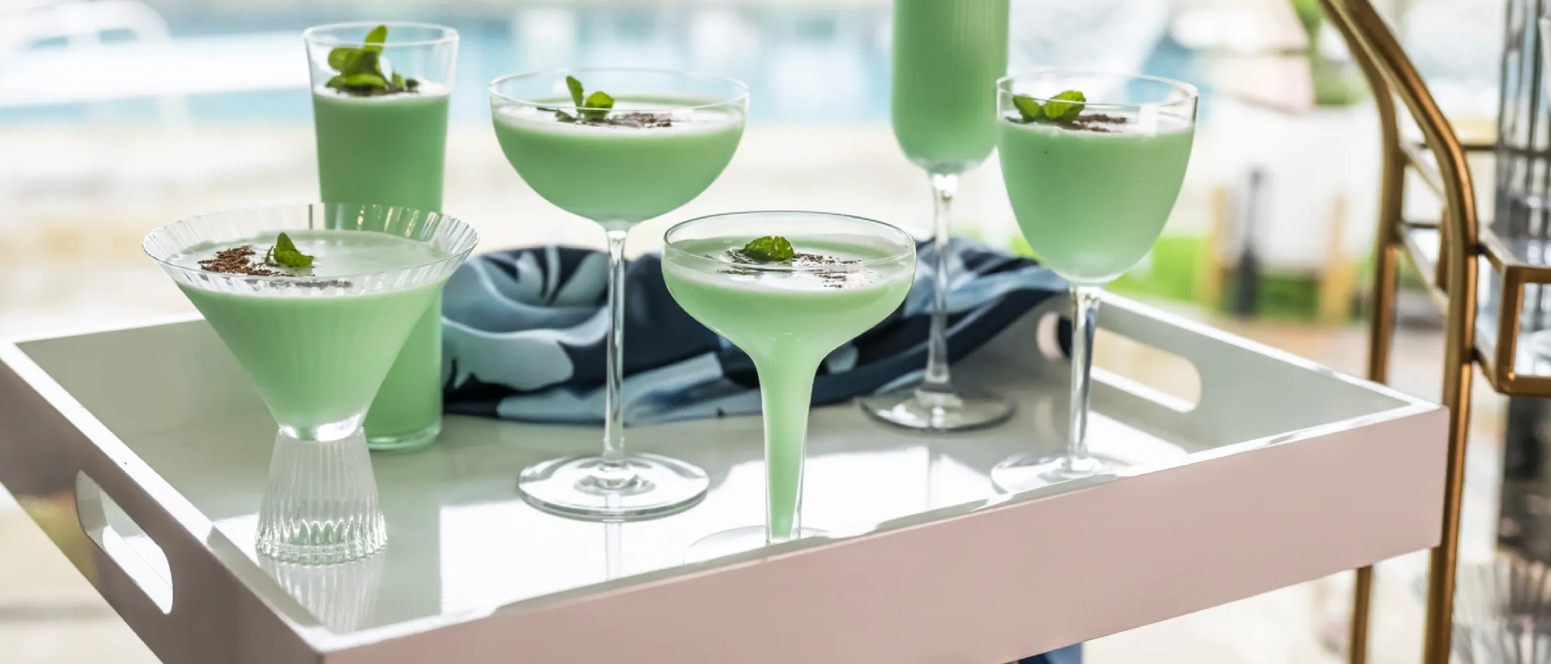 Six green mint cocktails in assorted glasses on a white tray by a poolside with a blue cloth