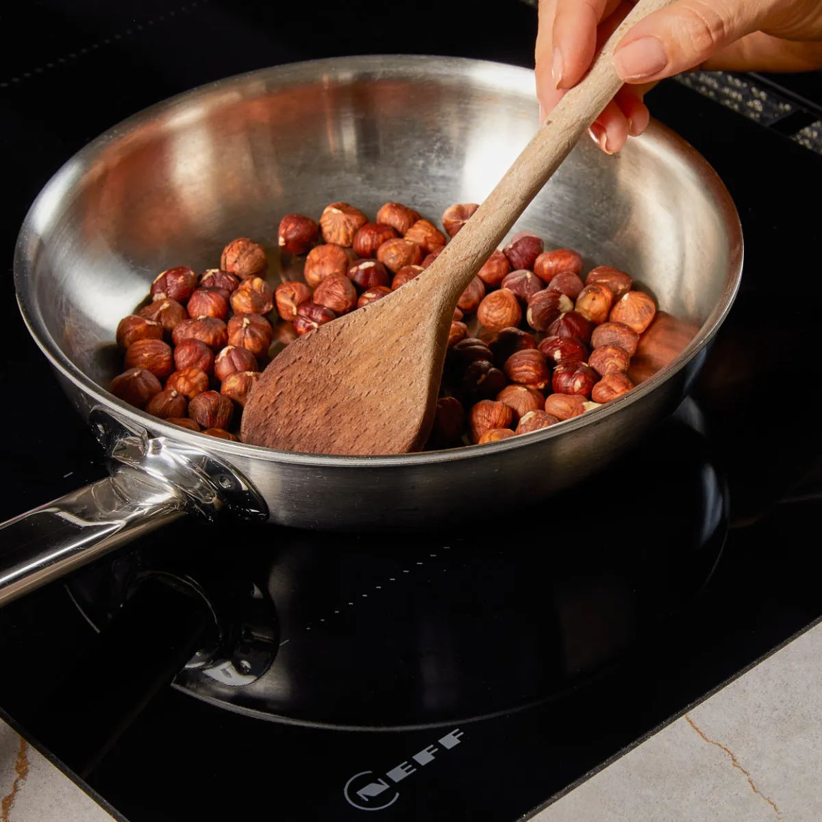 A hand stirring hazelnuts in a stainless steel pan on a black induction cooktop with a wooden spoon.