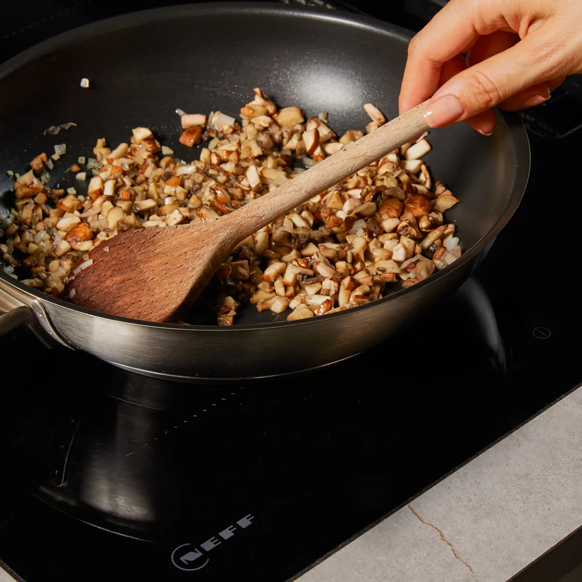 A hand stirs chopped mushrooms and onions in a pan on a black induction cooktop with a wooden spoon.
