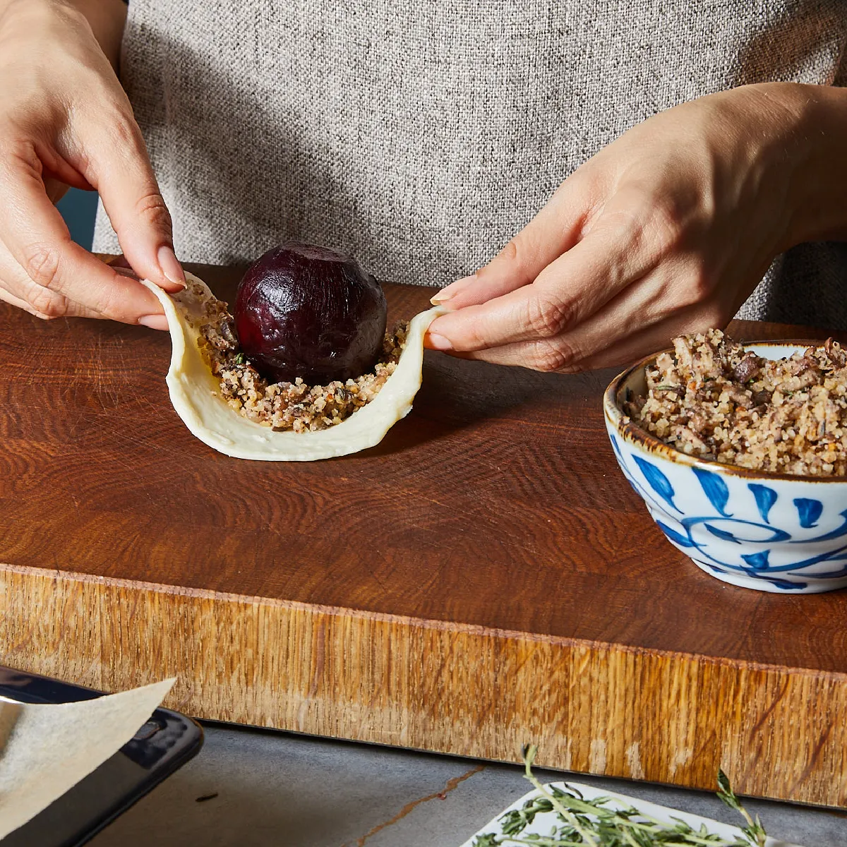 A person's hands are carefully wrapping a roasted beet and mushroom mixture in pastry dough on a wooden cutting board.