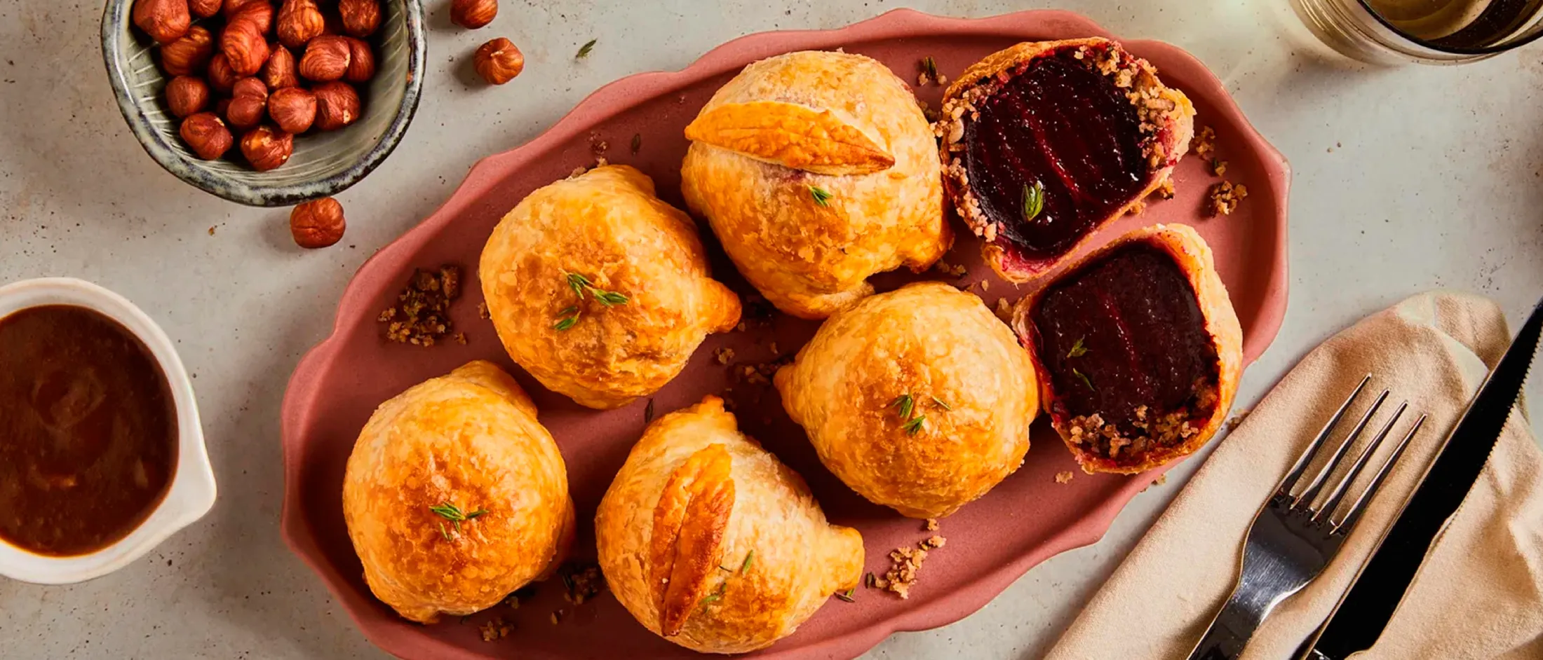 A pink platter holds golden-brown pastries, some whole and one cut open to reveal a dark filling. Hazelnuts and sauce are nearby.