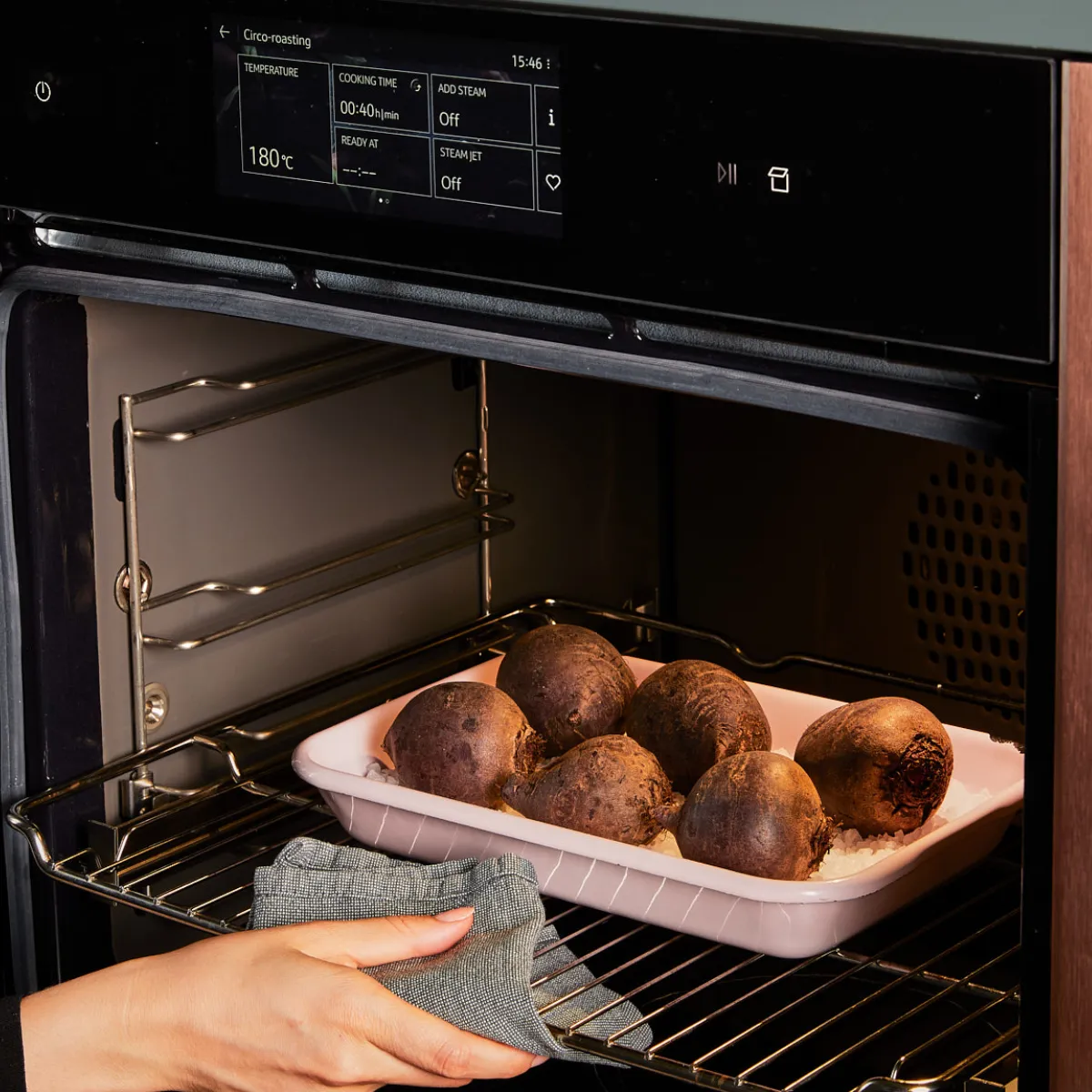 A person's hands, wearing a grey oven mitt, place a pink tray of roasted beets into a modern oven.
