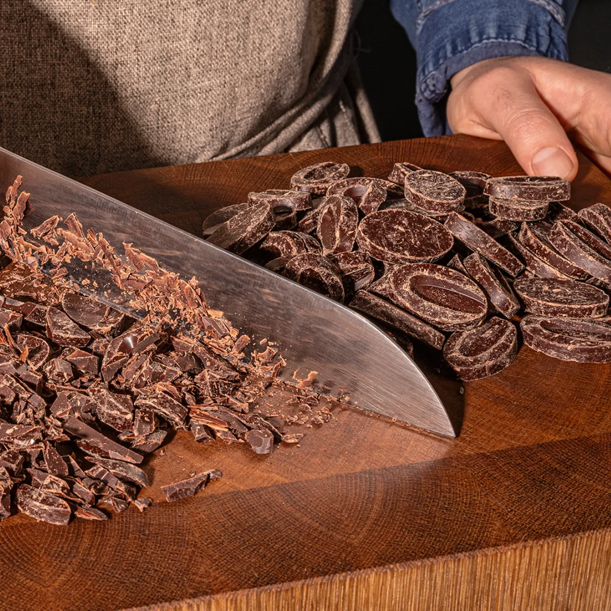 Hands chopping dark chocolate on a wooden cutting board.
