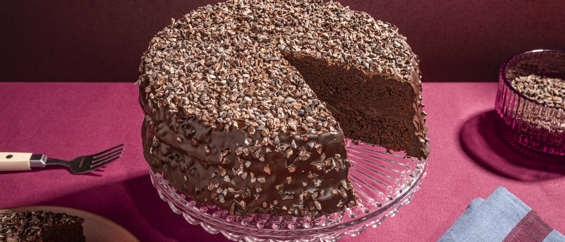 A rich chocolate cake, covered in chocolate chips, sits on a glass cake stand with a slice removed.