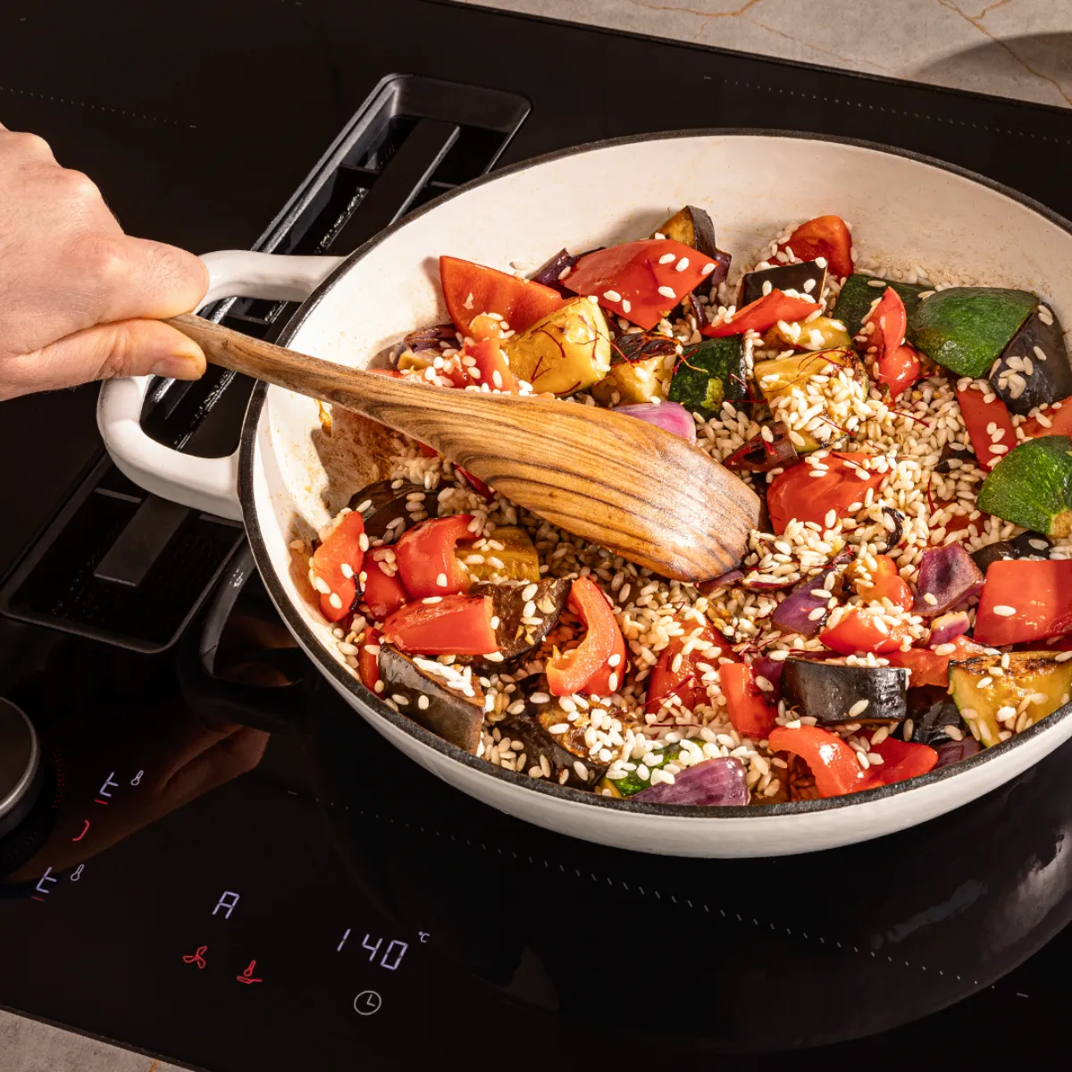 Cooking pan with healthy vegetable stir-fry, tomatoes, olives, and cooking utensils on stovetop.