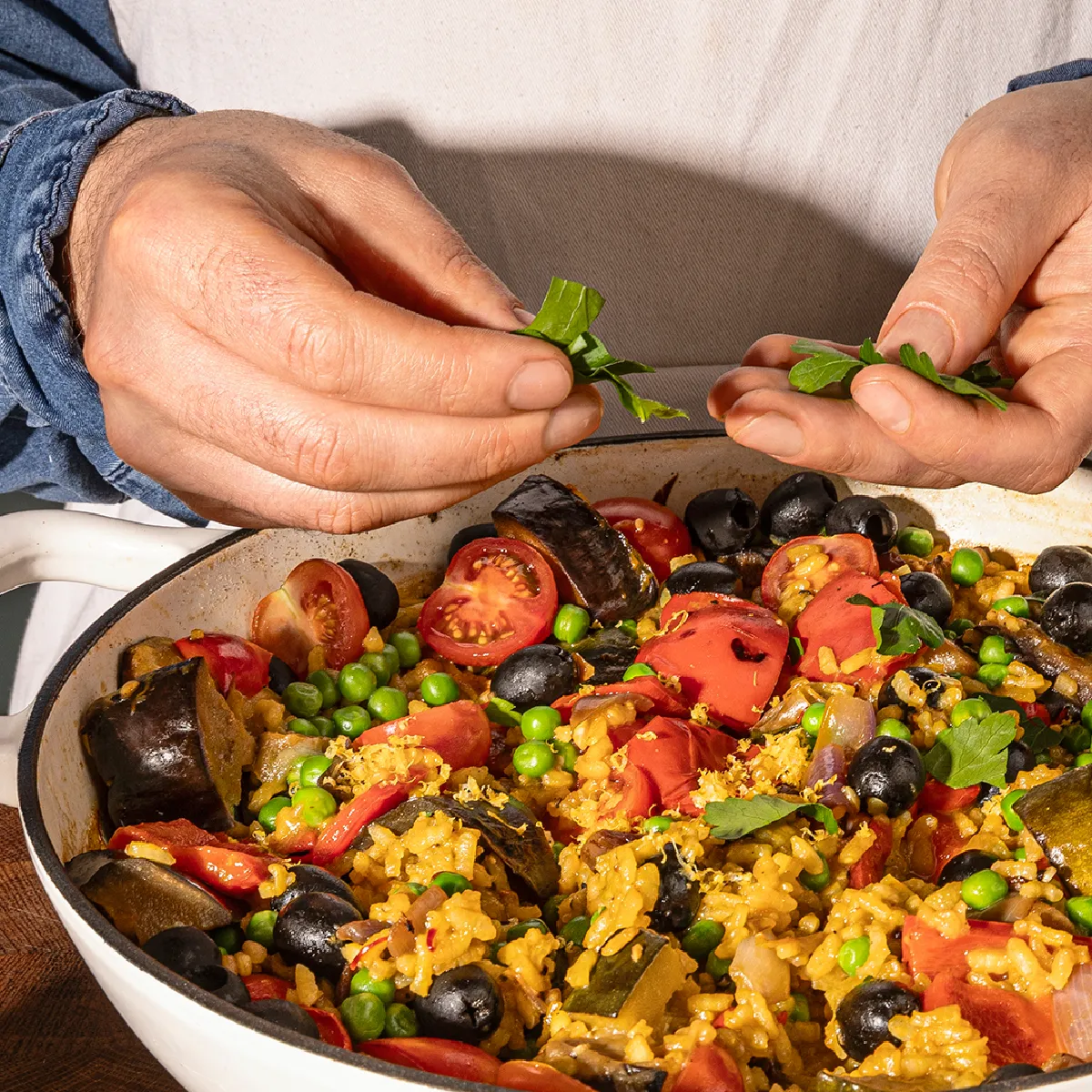 Hands garnishing a pan of colorful vegetable and rice dish with fresh herbs.