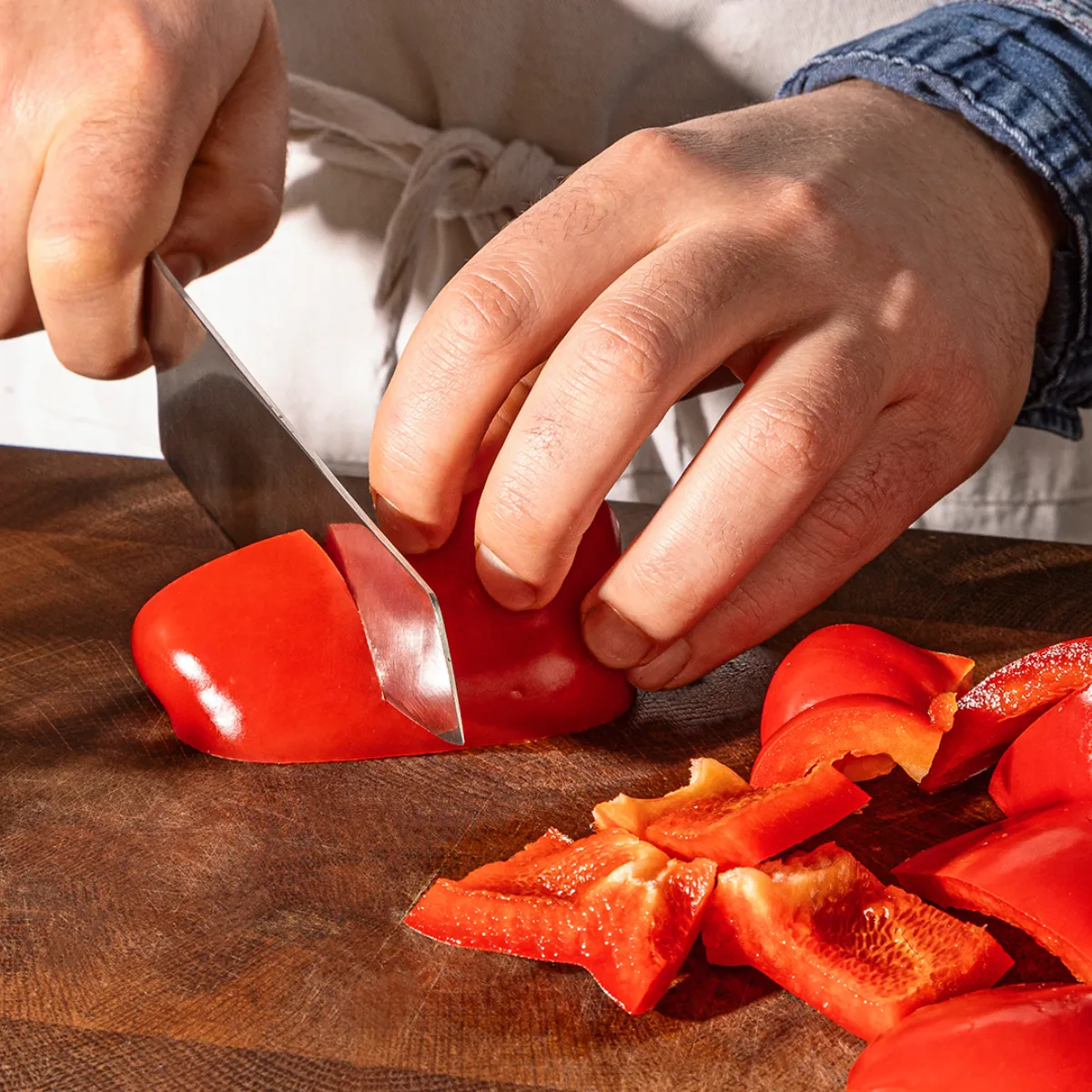 Hands chopping red bell peppers on a wooden cutting board, with fresh herbs in the background.