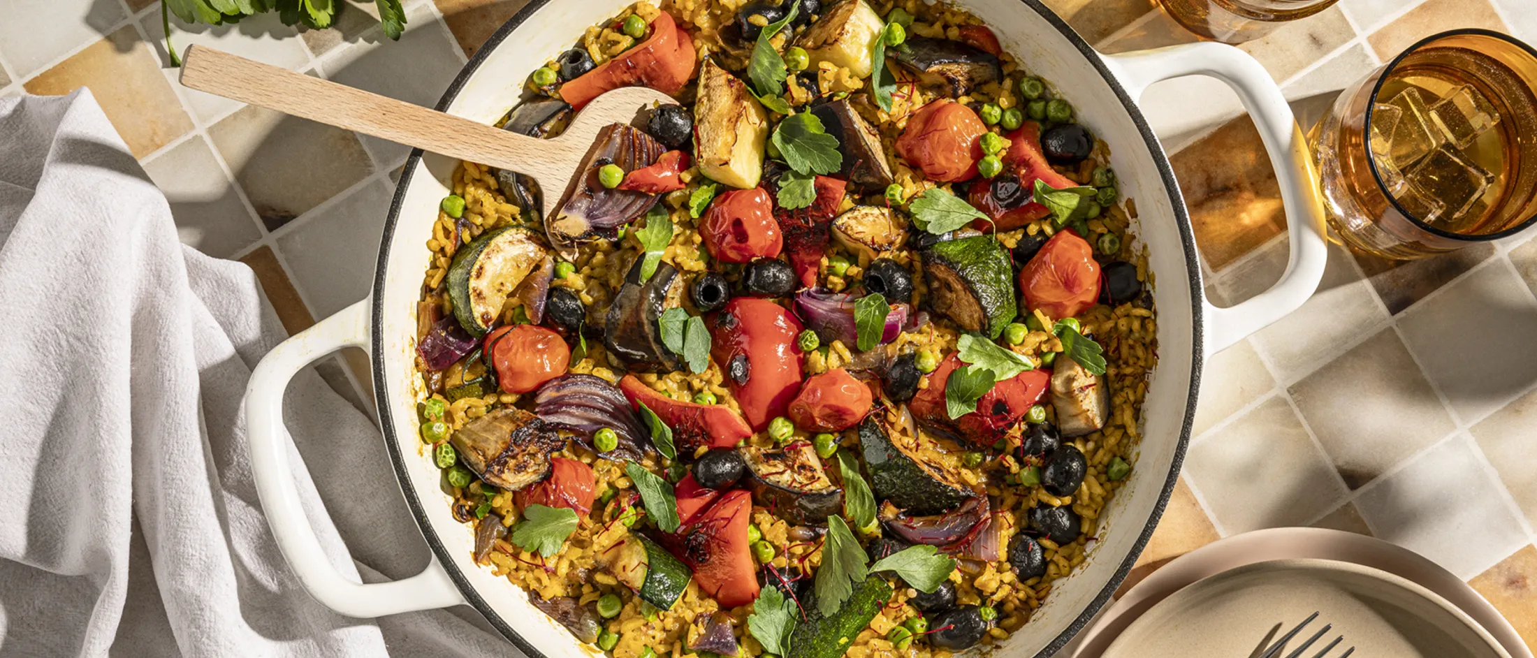 A large white pan filled with colorful paella, featuring roasted vegetables, rice, and herbs, with a wooden spoon.