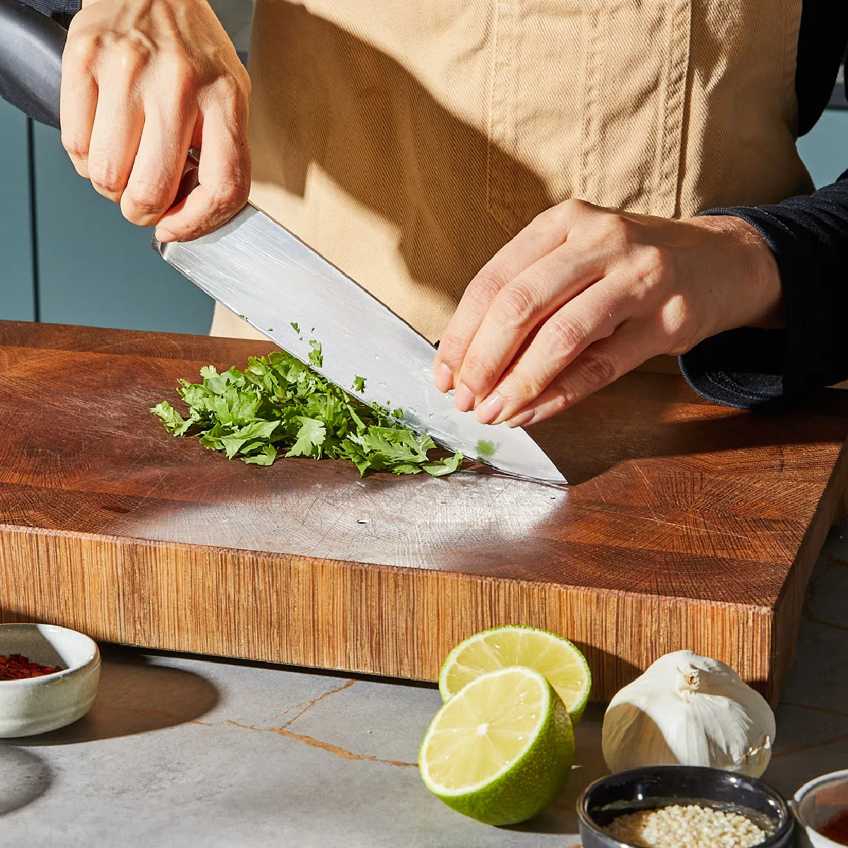 Auf einem Holzschneidebrett hackt jemand mit einem großen Messer frischen Koriander, daneben liegen Limetten und Knoblauch.