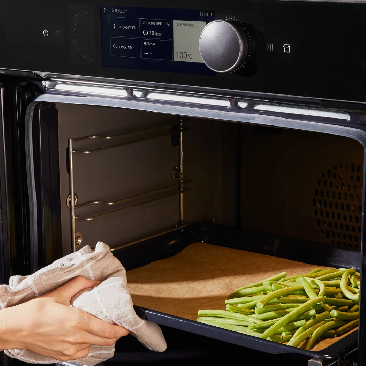 Hands placing a tray of green beans into a modern oven with a digital display showing cooking settings.