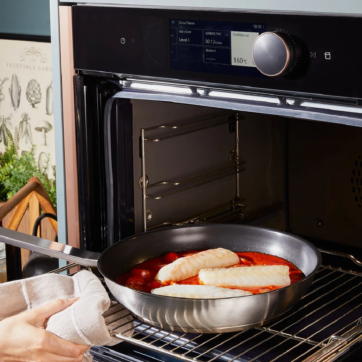 Hand placing a frying pan with fish fillets and tomato sauce into a modern oven set at 160°C with digital display.