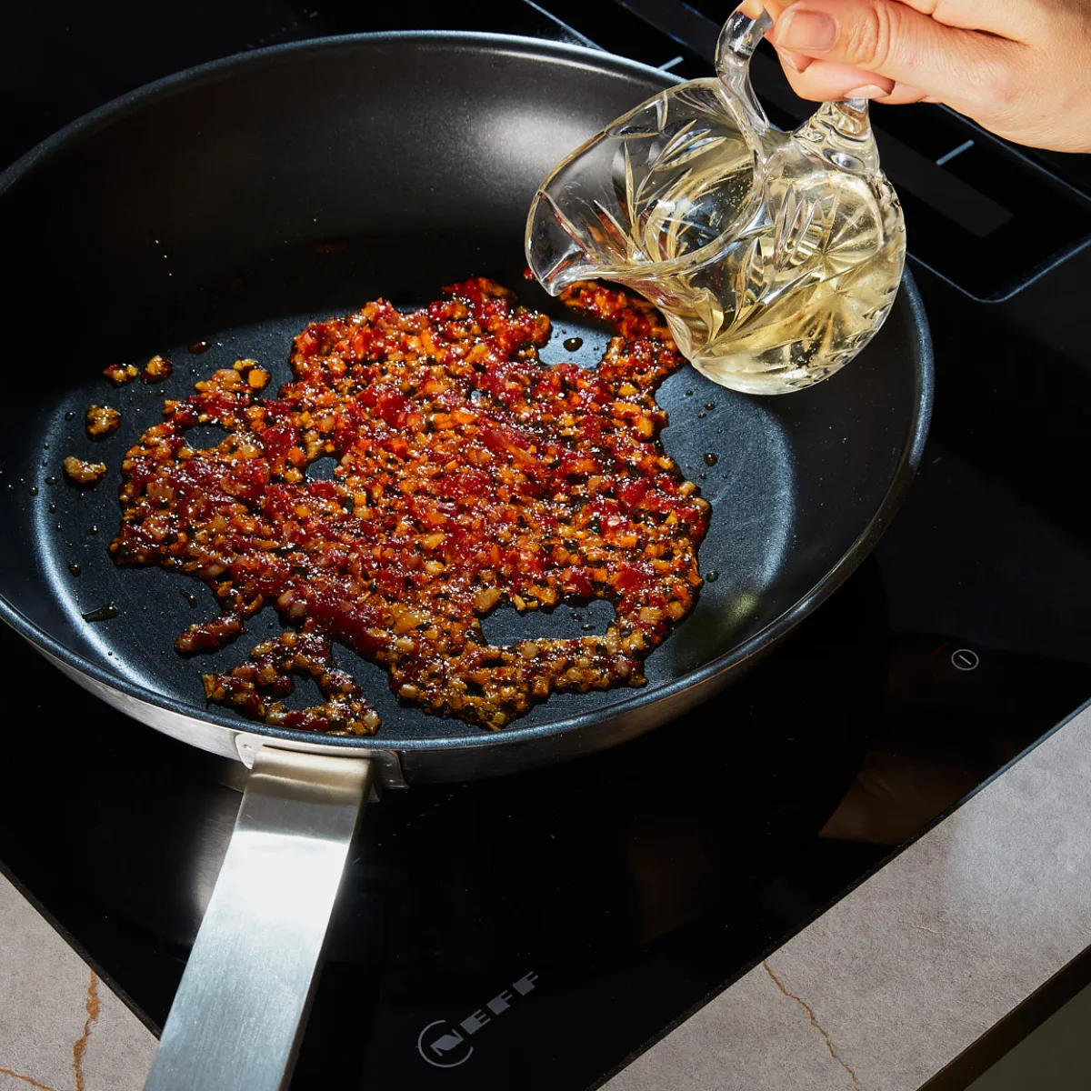 Hand pouring oil into a pan with sautéed minced garlic and chili flakes on an electric stove.