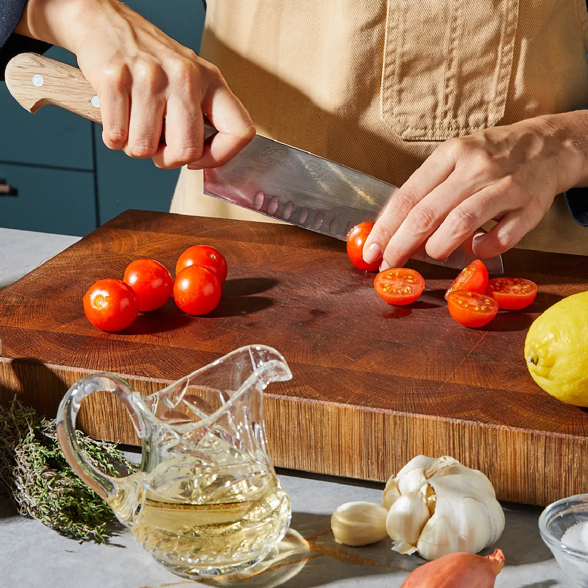 Person slicing cherry tomatoes on a wooden cutting board with garlic, thyme, lemon, and oil nearby.