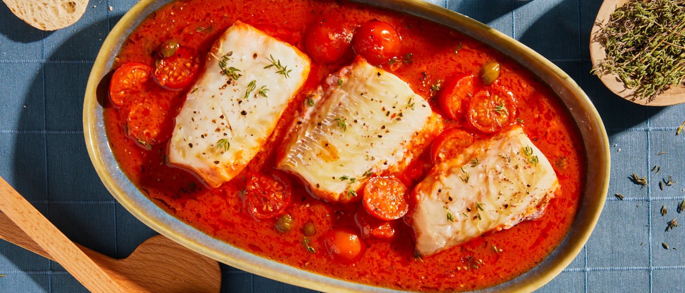 Baked white fish fillets in a tomato-based sauce with cherry tomatoes and herbs, served in an oval ceramic dish.