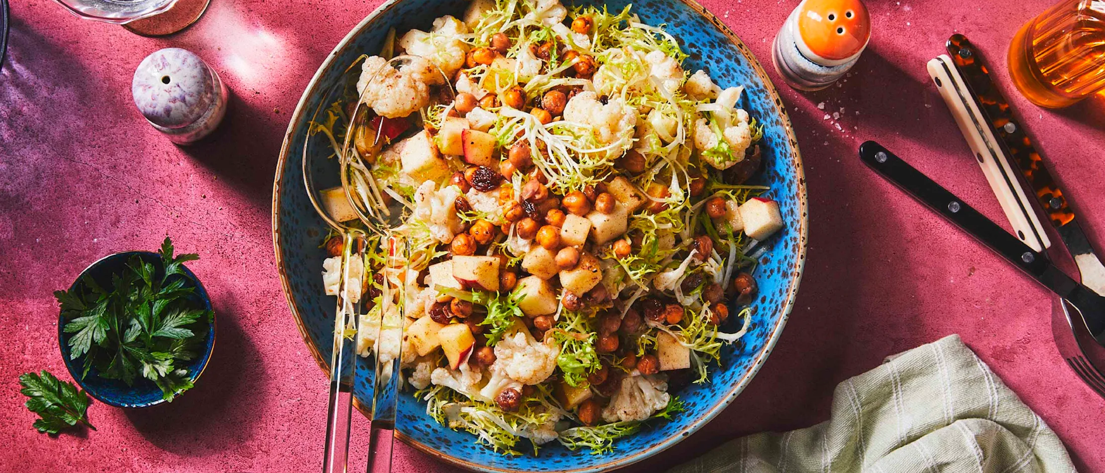 A big blue bowl filled with cauliflower salad surrounded by colorful dinner table dishes, like a fork, a napkin, plates and a glas filled with water.