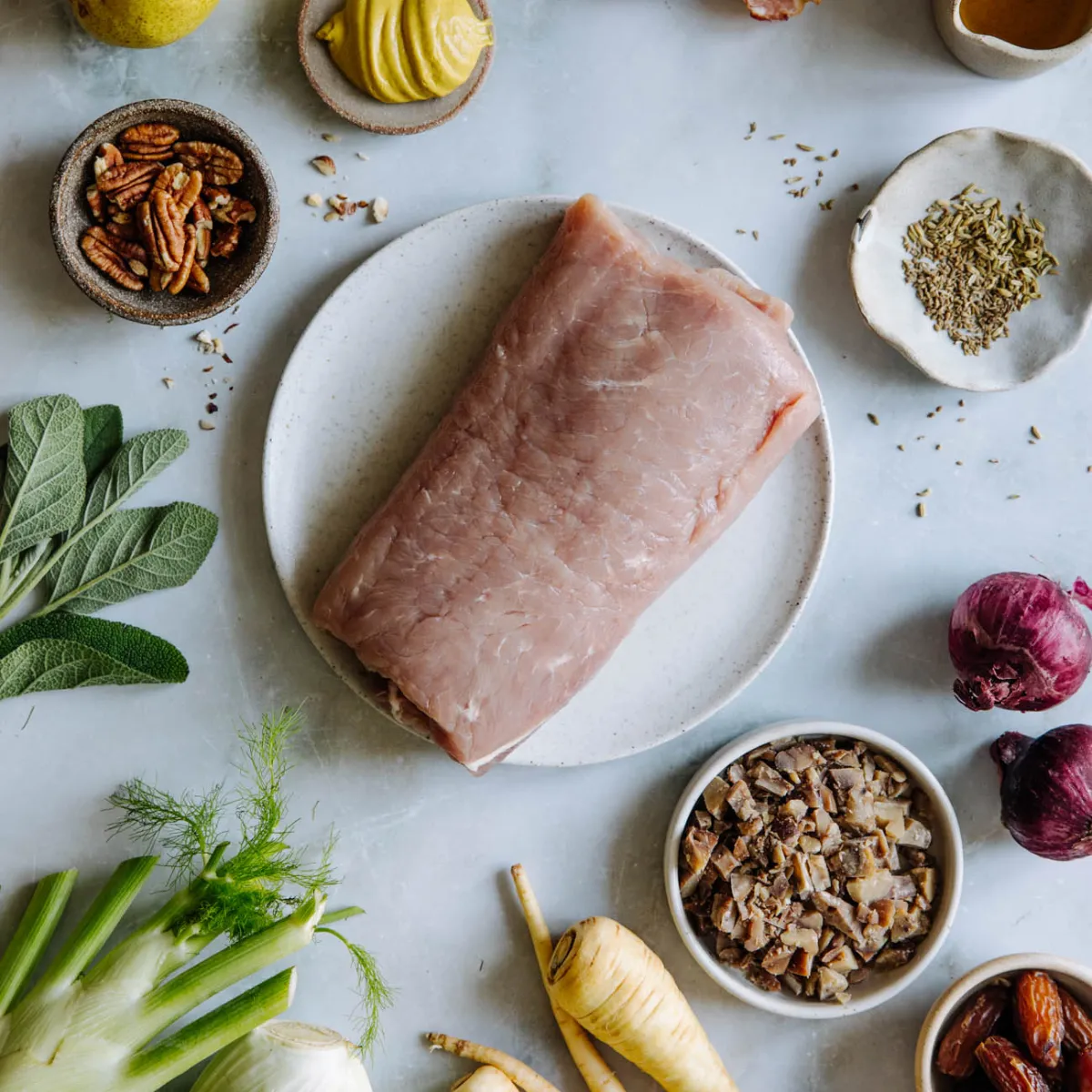Rohes Schweinefleisch auf einem Teller, umgeben von Kräutern, Nüssen, gehackten Pilzen, Zwiebeln und Gemüse auf einem hellen Untergrund.