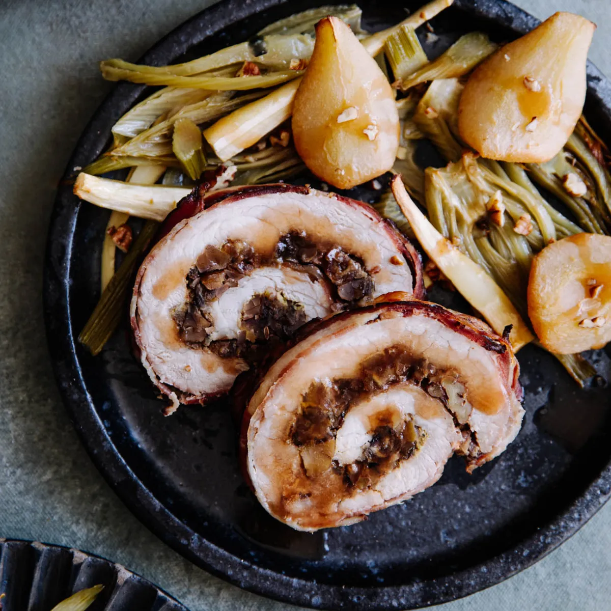 In Scheiben geschnittener Schweinebraten mit Füllung, serviert mit geschmortem Lauch und Birnen auf einem dunklen Teller.