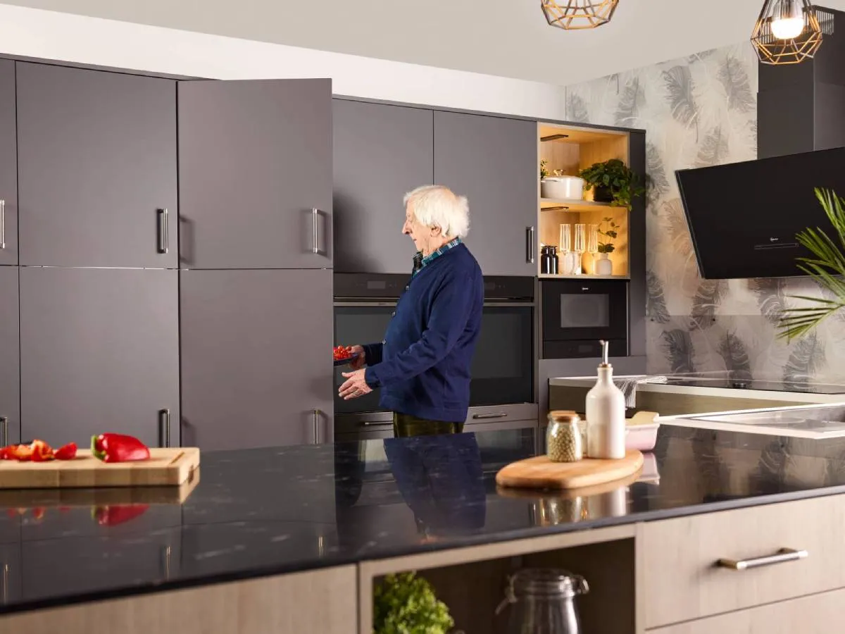 An elderly gentleman standing by grey kitchen cabinets, opening a built-in fridge door and built‑in ovens holding a bowl of cherry tomatoes