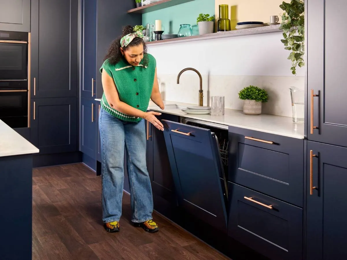 Woman in a green sleeveless knitted jumper and headband stands in a navy-blue kitchen, opening a built-in dishwasher and reaching toward the interior of the machine