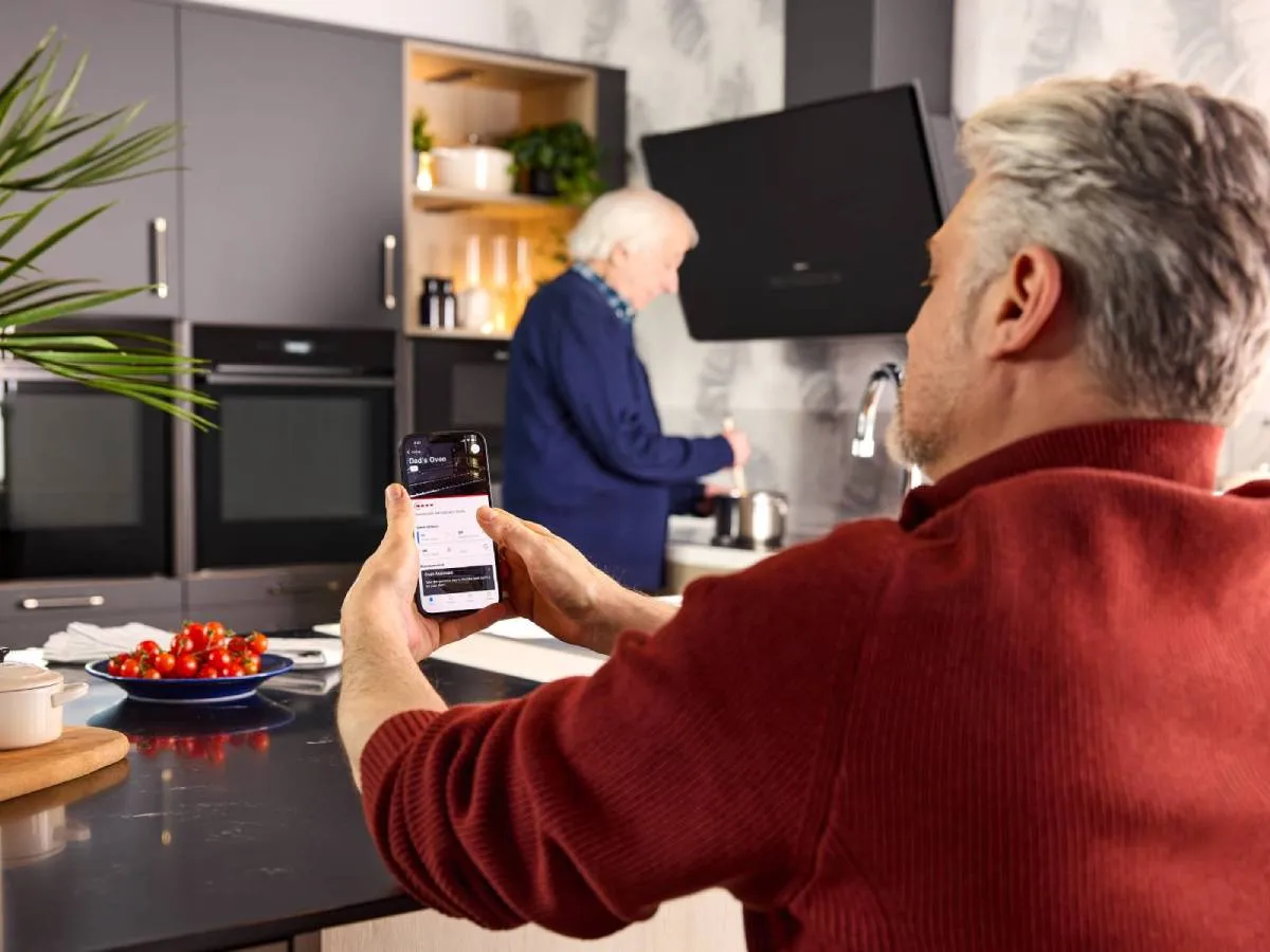 An elderly person cooks at the hob. His son monitors the Home Connect app remotely for added safety and peace of mind.