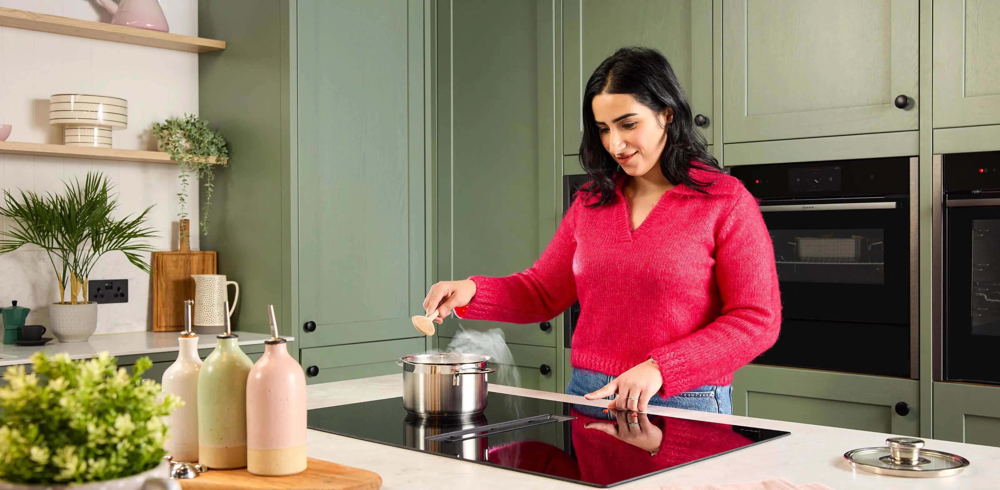 In a green kitchen, a woman with low vision stands at an induction hob. She uses tactile markers on touch controls for safe, precise control.
