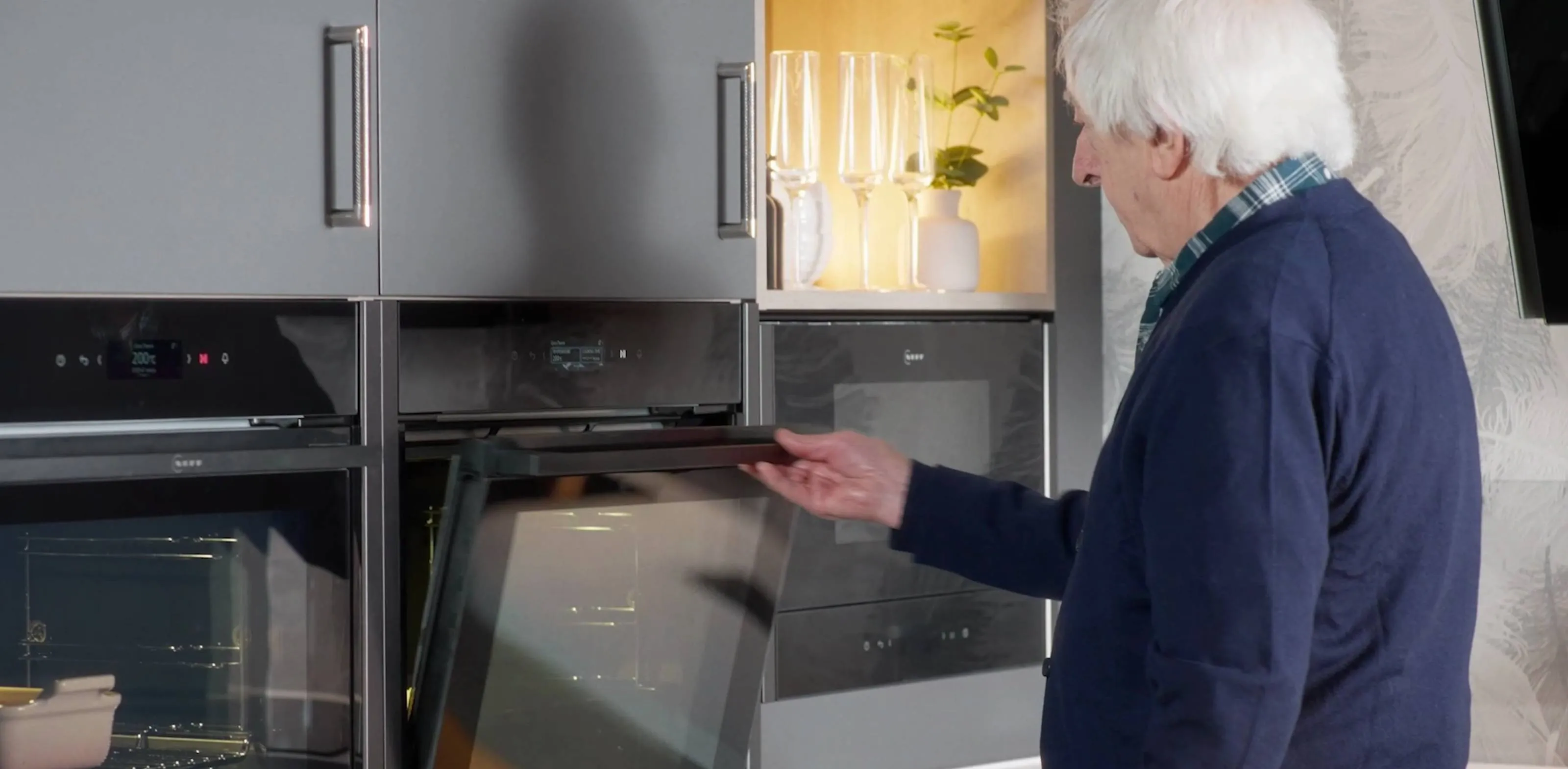 An elderly gentleman standing by grey kitchen cabinets, opening a built-in slide and hide oven door.