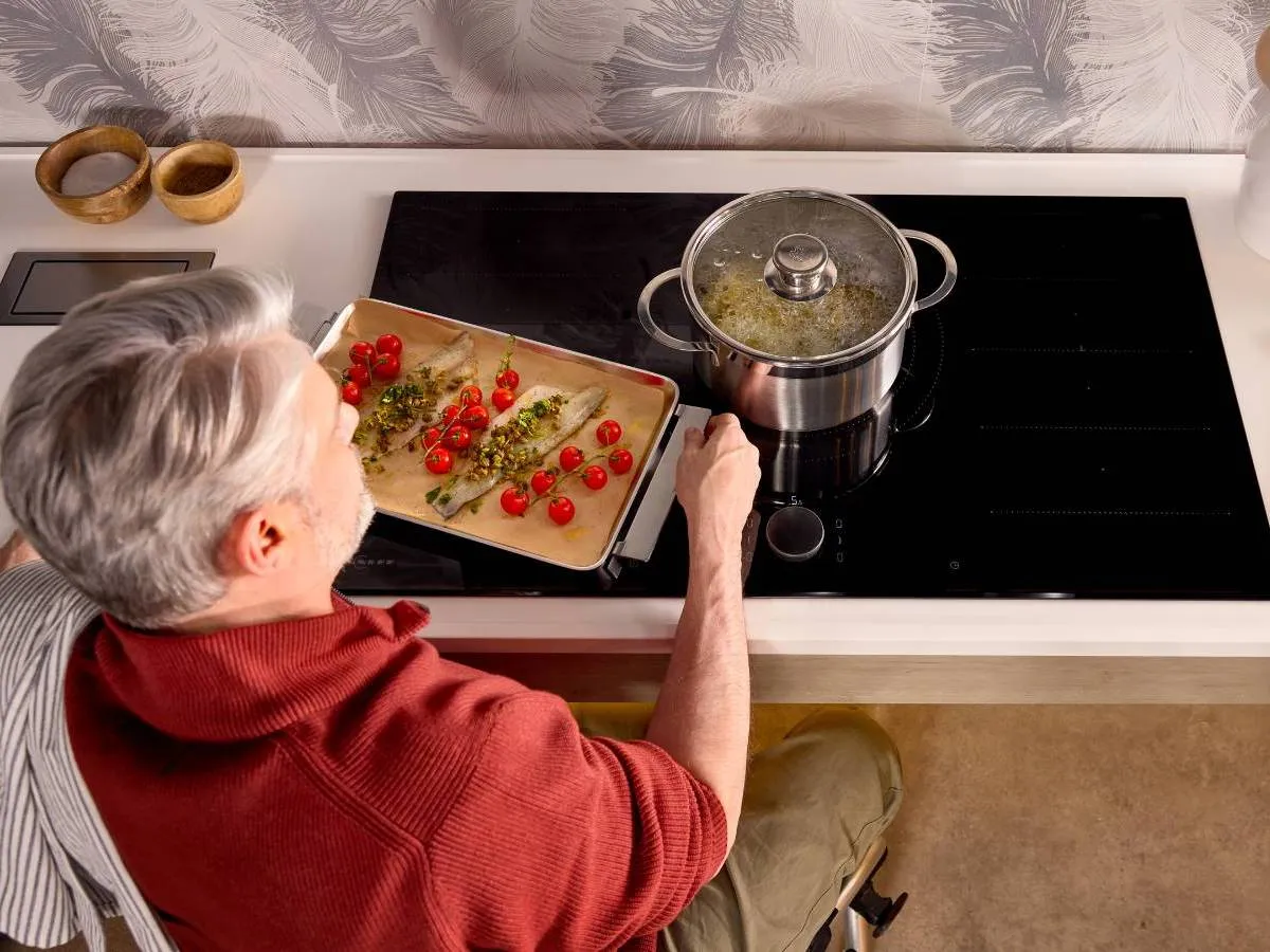 Seen from overhead, a wheelchair user manoeuvres under an induction hob. Tray of fish and vegetables moves easily across the flat hob surface.
