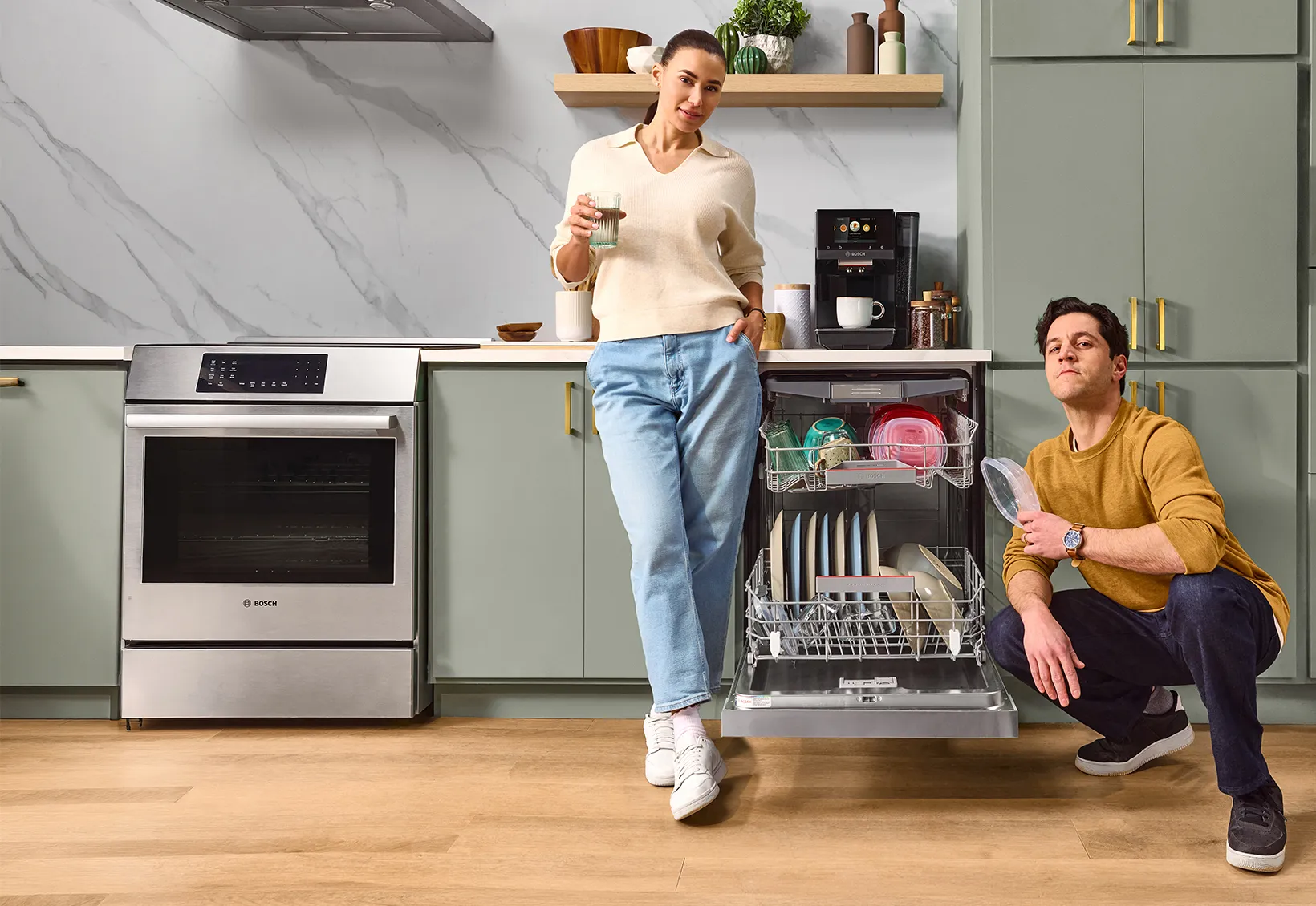 A woman and a man in a modern kitchen beside an open dishwasher, the woman holding a cup and the man squatting on the floor.