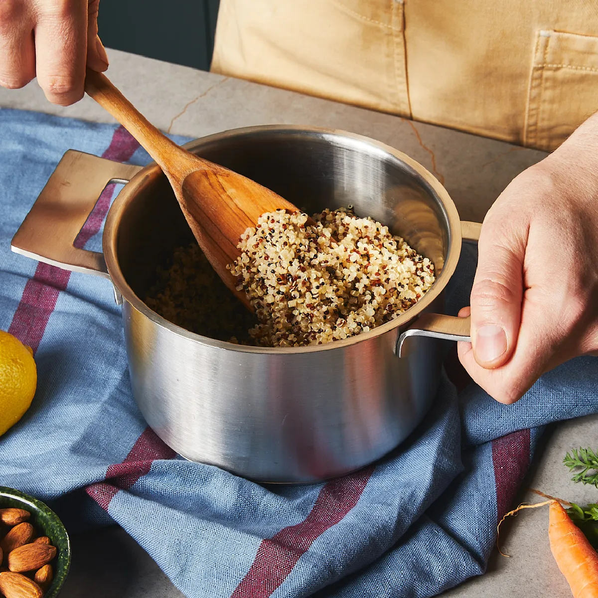 Hands stirring cooked quinoa with a wooden spoon in a stainless steel pot on a blue cloth with almonds and a carrot nearby.