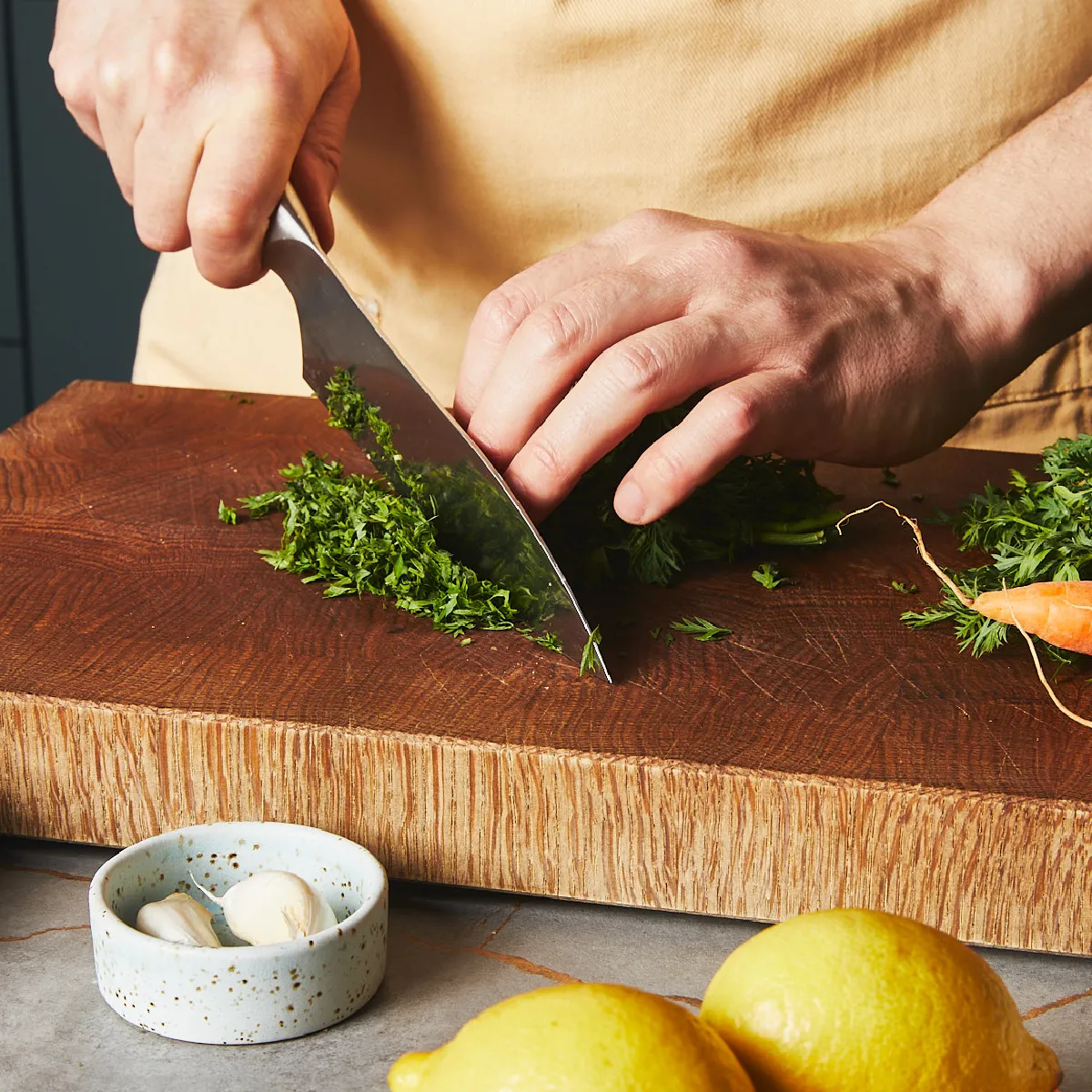 Hands chopping fresh green herbs on a wooden cutting board, with garlic cloves and lemons nearby.