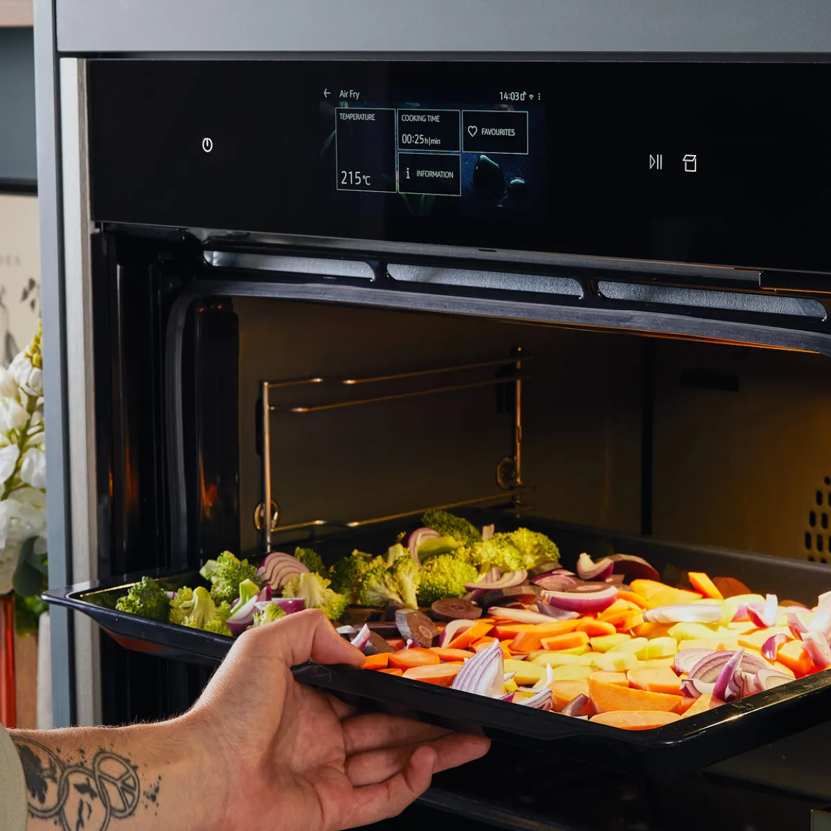 Hand with tattoo placing a tray of chopped vegetables into a modern digital oven set to air fry at 215°C for 25 minutes.