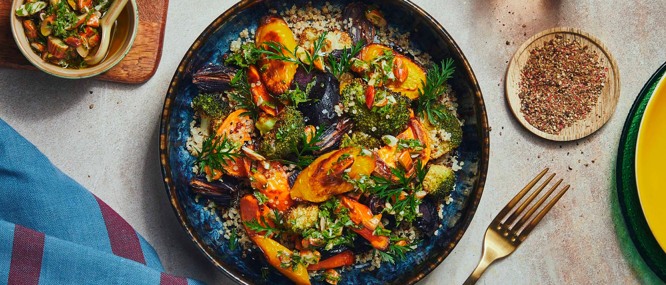 A bowl of roasted mixed vegetables with quinoa, garnished with herbs, a small bowl of dressing, pepper flakes, and a gold fork nearby.