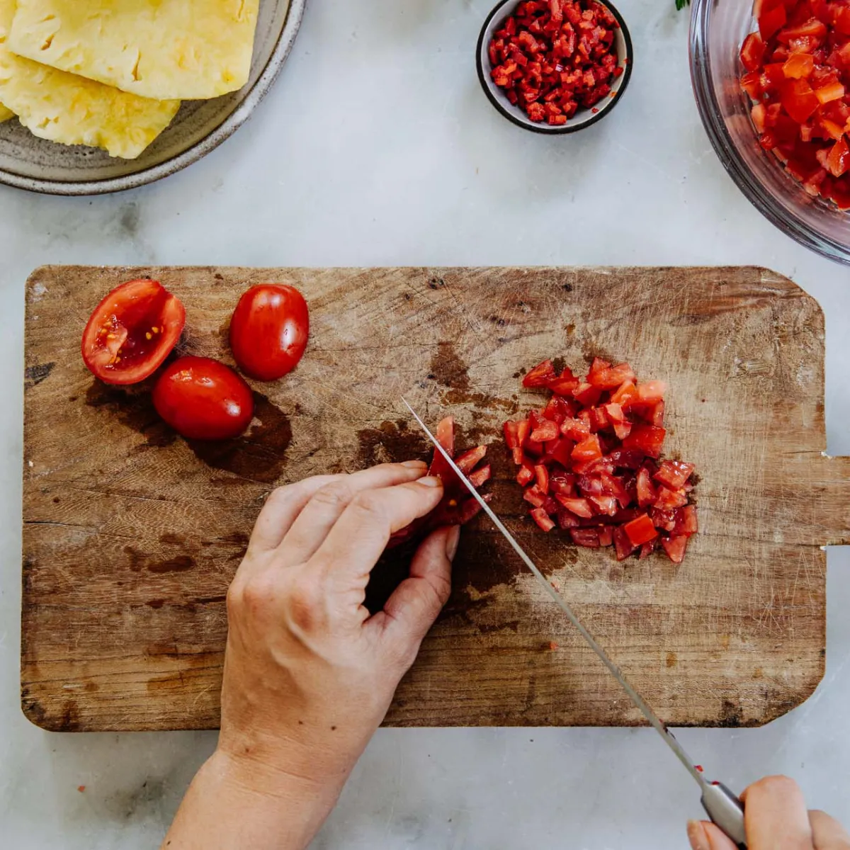 Hands chopping tomatoes on a wooden cutting board with pineapple slices, chopped onions, chili, and cilantro nearby on a kitchen countertop.