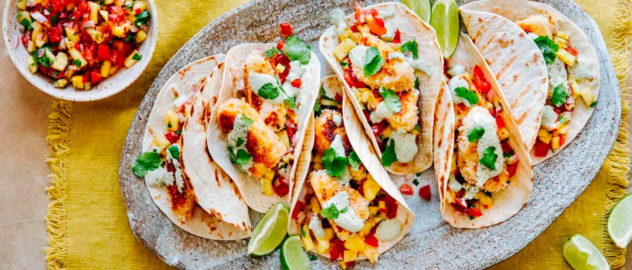 Grilled fish tacos with creamy sauce, fresh pineapple salsa, cilantro, and lime wedges on a stone platter over a yellow cloth. 