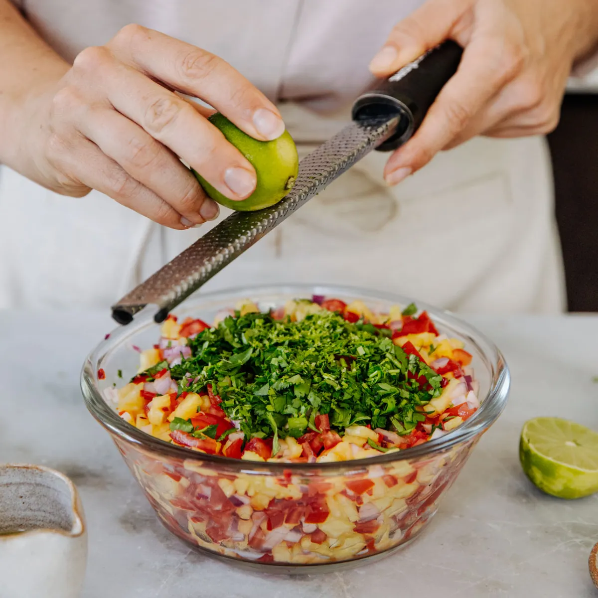 Woman zesting a fresh lime directly into a vibrant bowl of salsa, adding a citrusy finish to the dish.