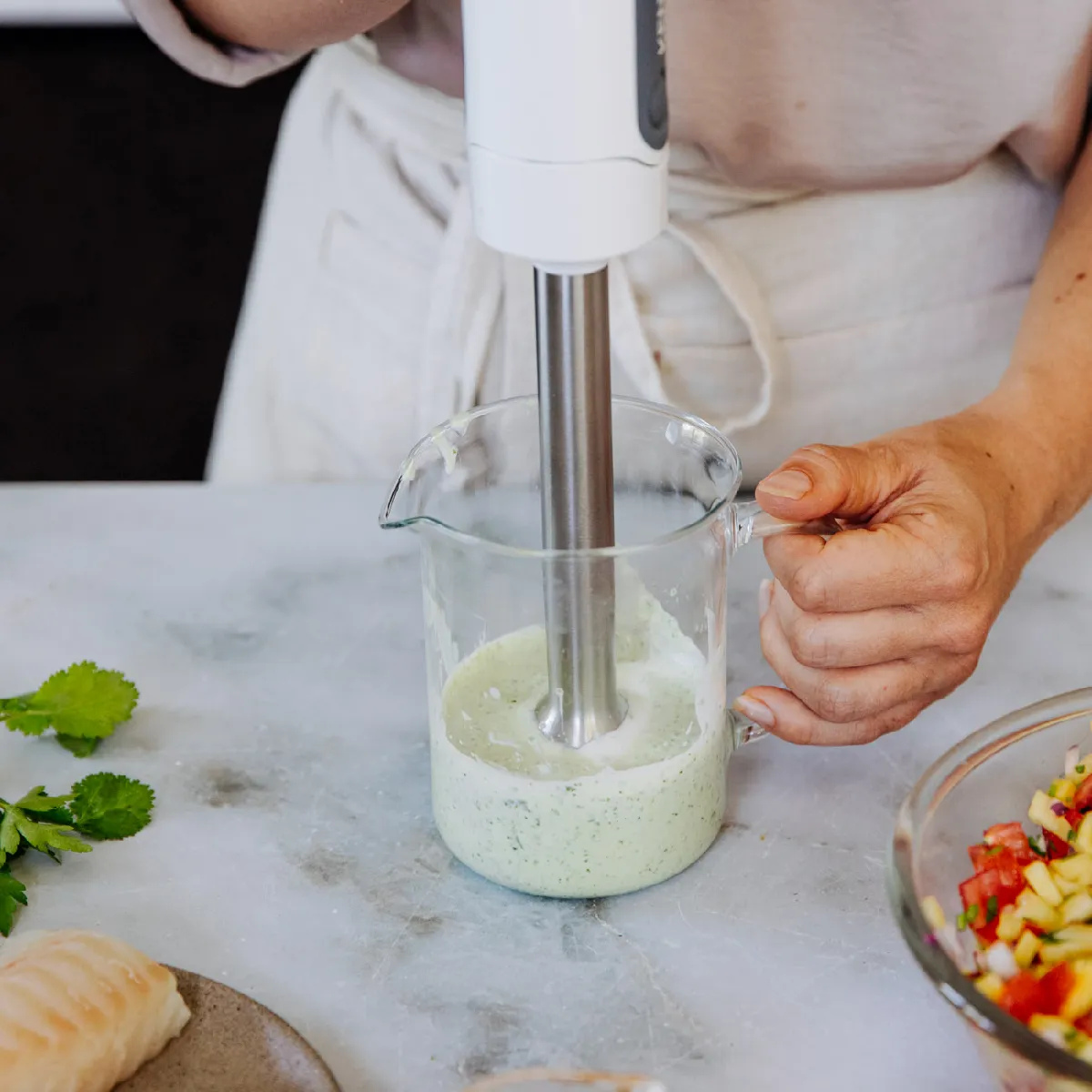 A person uses an immersion blender to mix a light green sauce in a clear measuring cup on a marble counter.