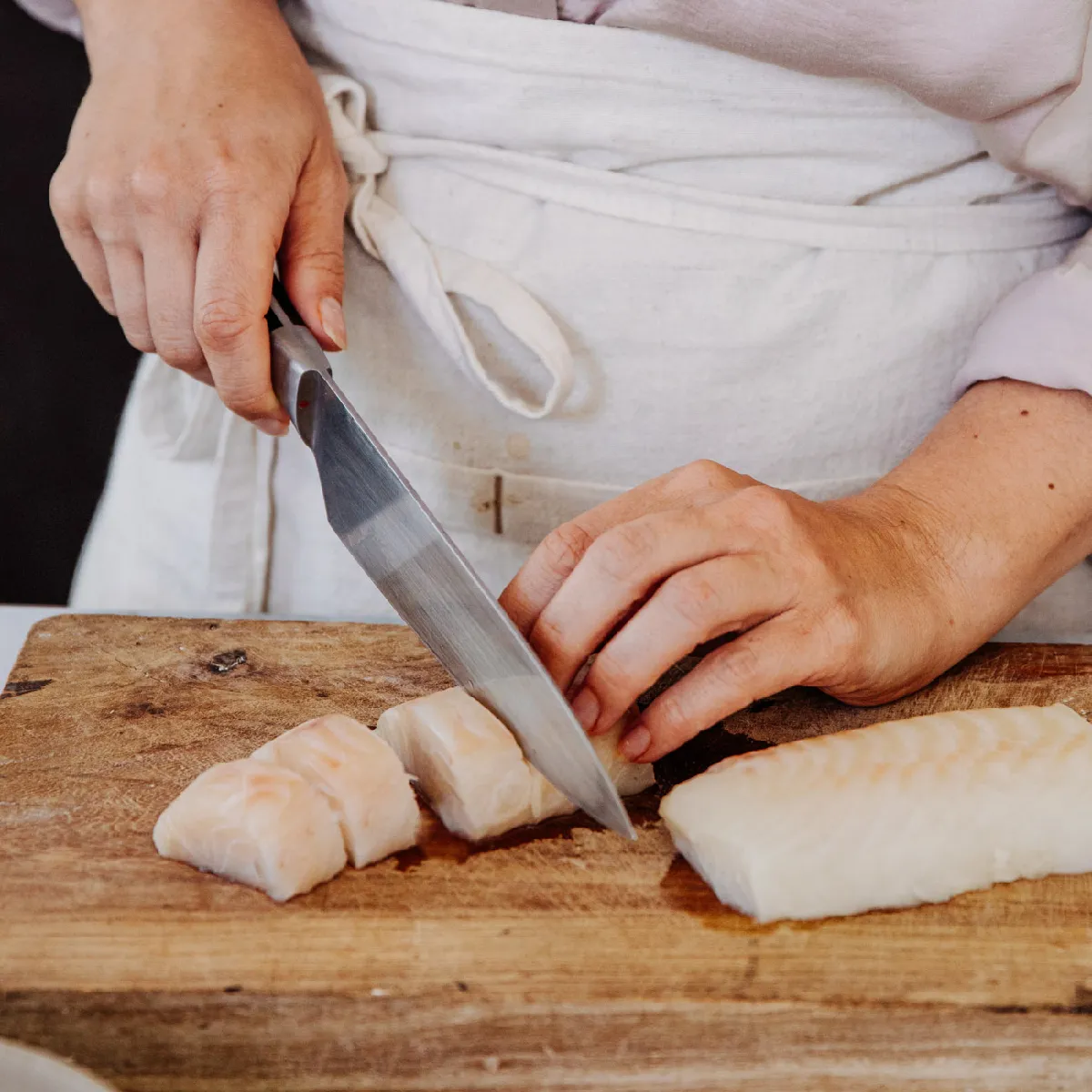 A person in an apron carefully slices raw fish fillets on a rustic wooden cutting board with a large knife.
