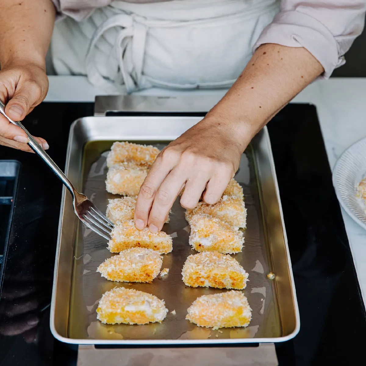 A person arranges breaded fish pieces on a baking tray, preparing them for cooking.