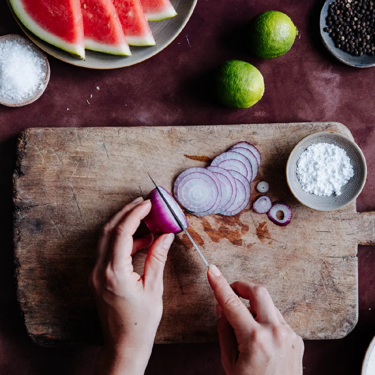 Overhead shot of hands thinly slicing a red onion on a rustic wooden cutting board, surrounded by ingredients.