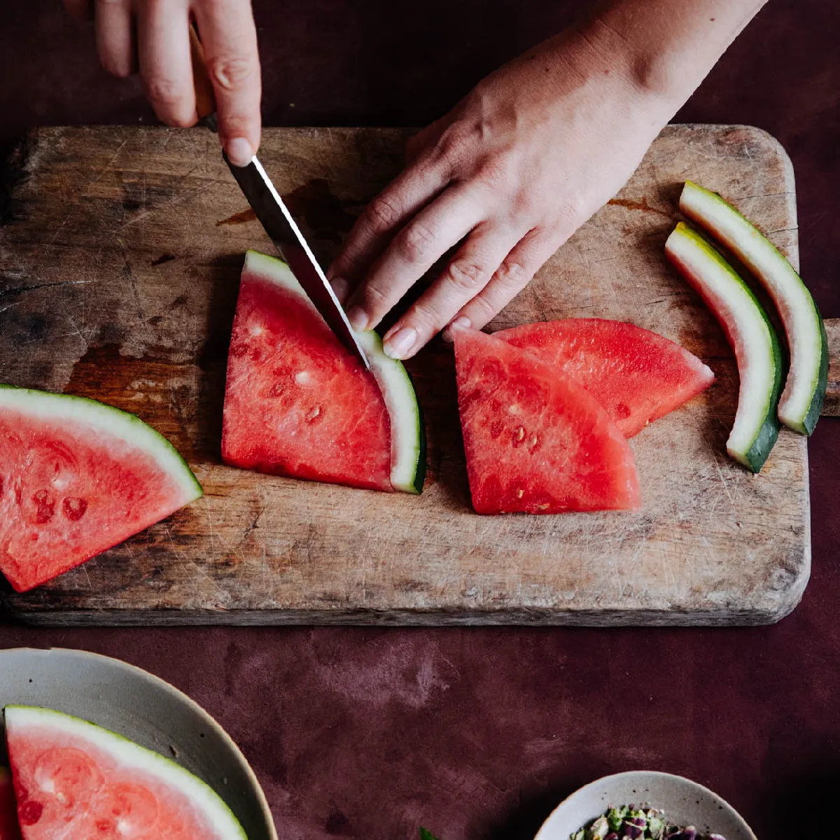 Hands carefully trim the rind from a slice of watermelon on a rustic wooden cutting board, with other slices nearby.