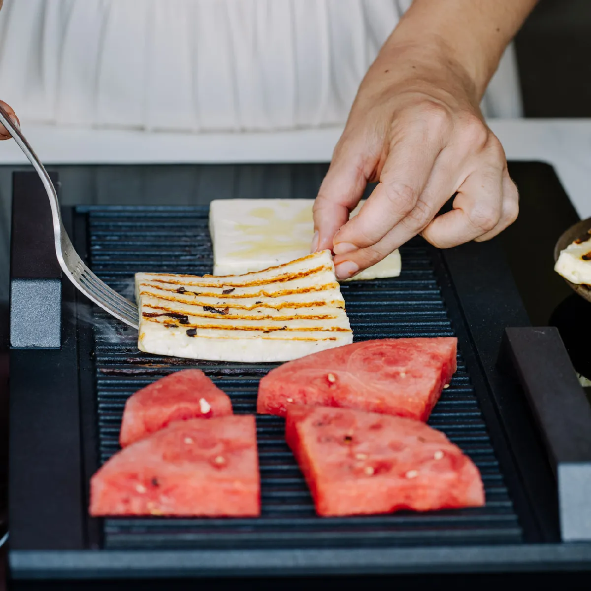 A person uses a fork to flip a piece of halloumi cheese on a grill, alongside watermelon slices.