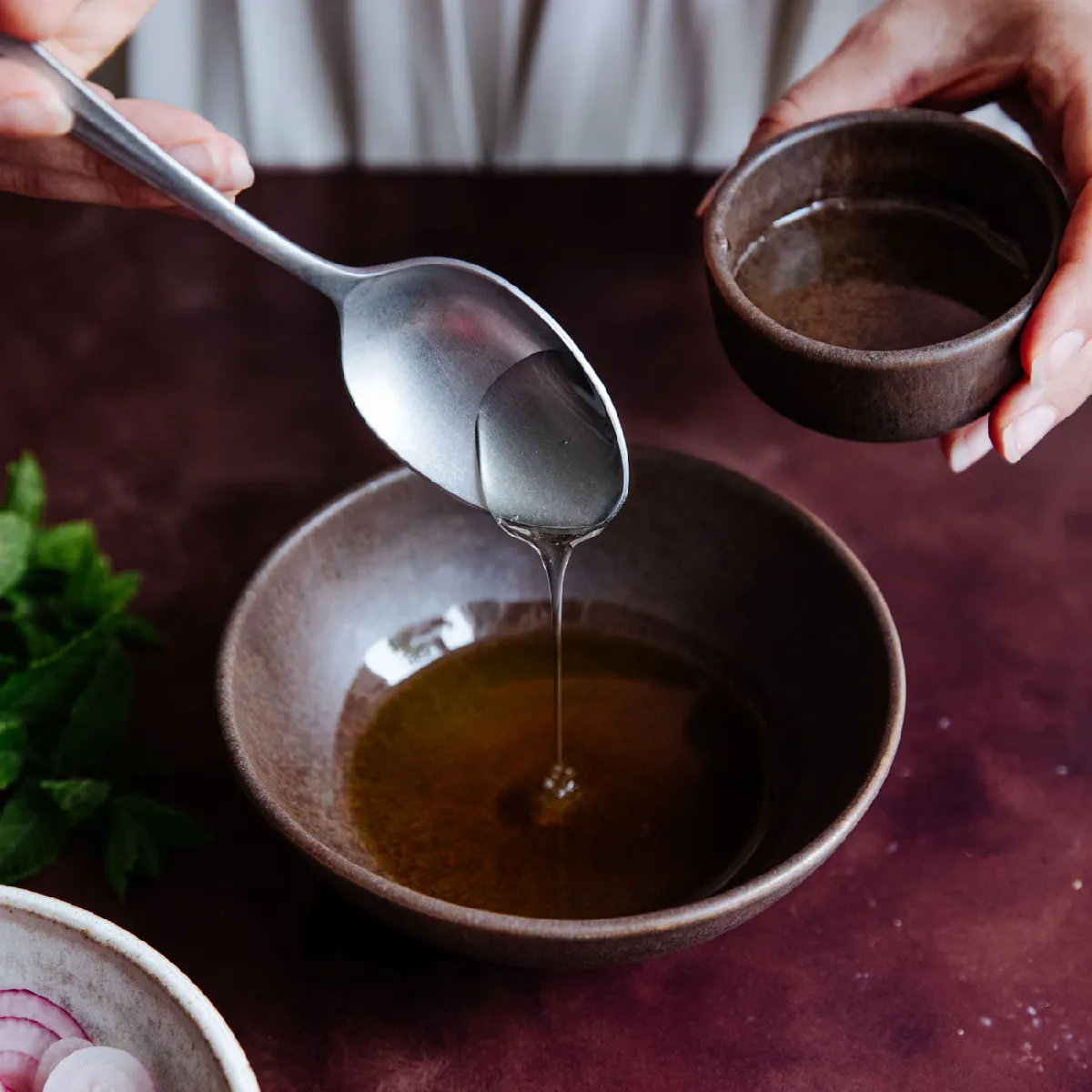 Hands pour a clear liquid from a spoon into a small brown bowl containing a darker liquid, likely oil.