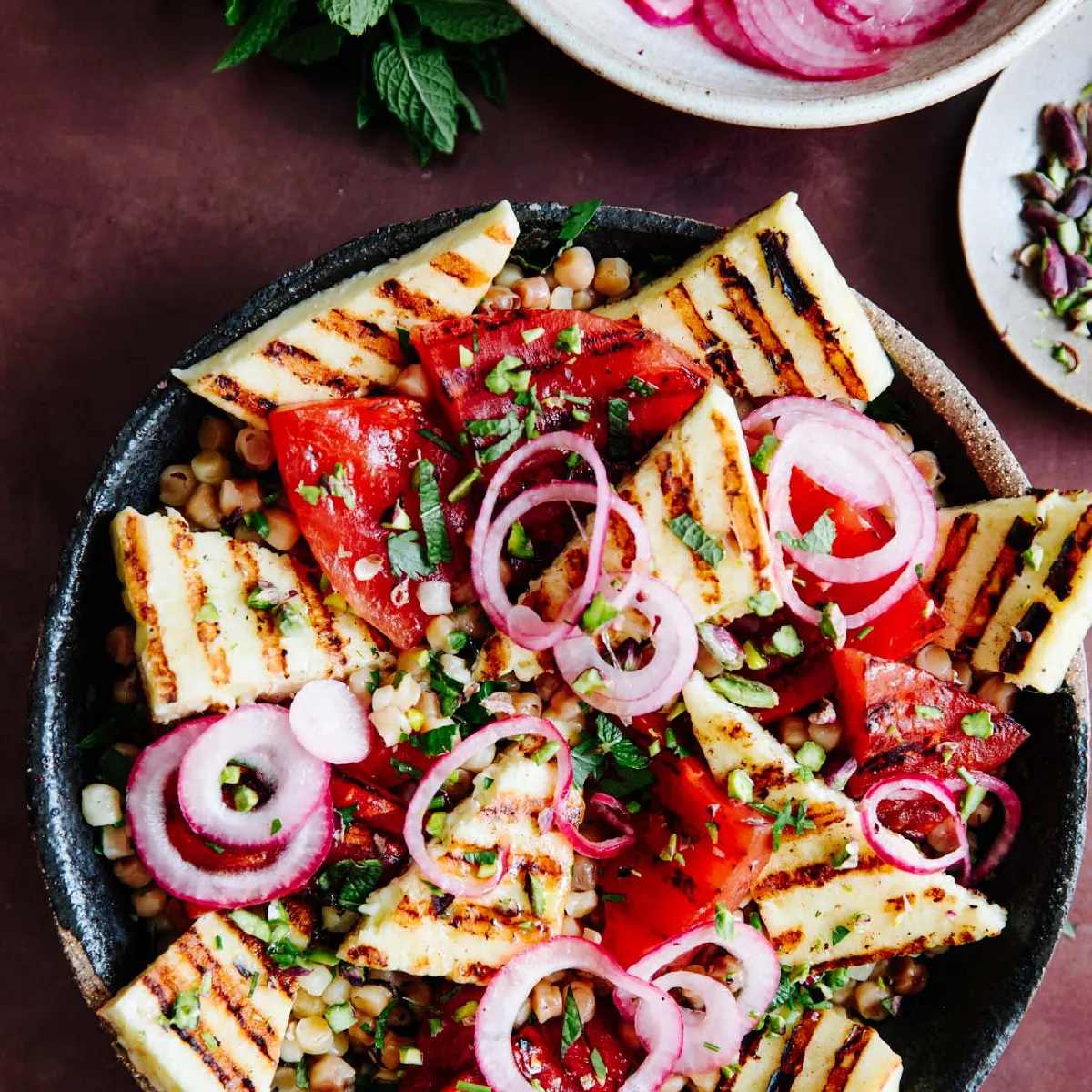 A close-up of a vibrant salad with grilled halloumi, tomatoes, chickpeas, red onion, and herbs in a dark bowl.