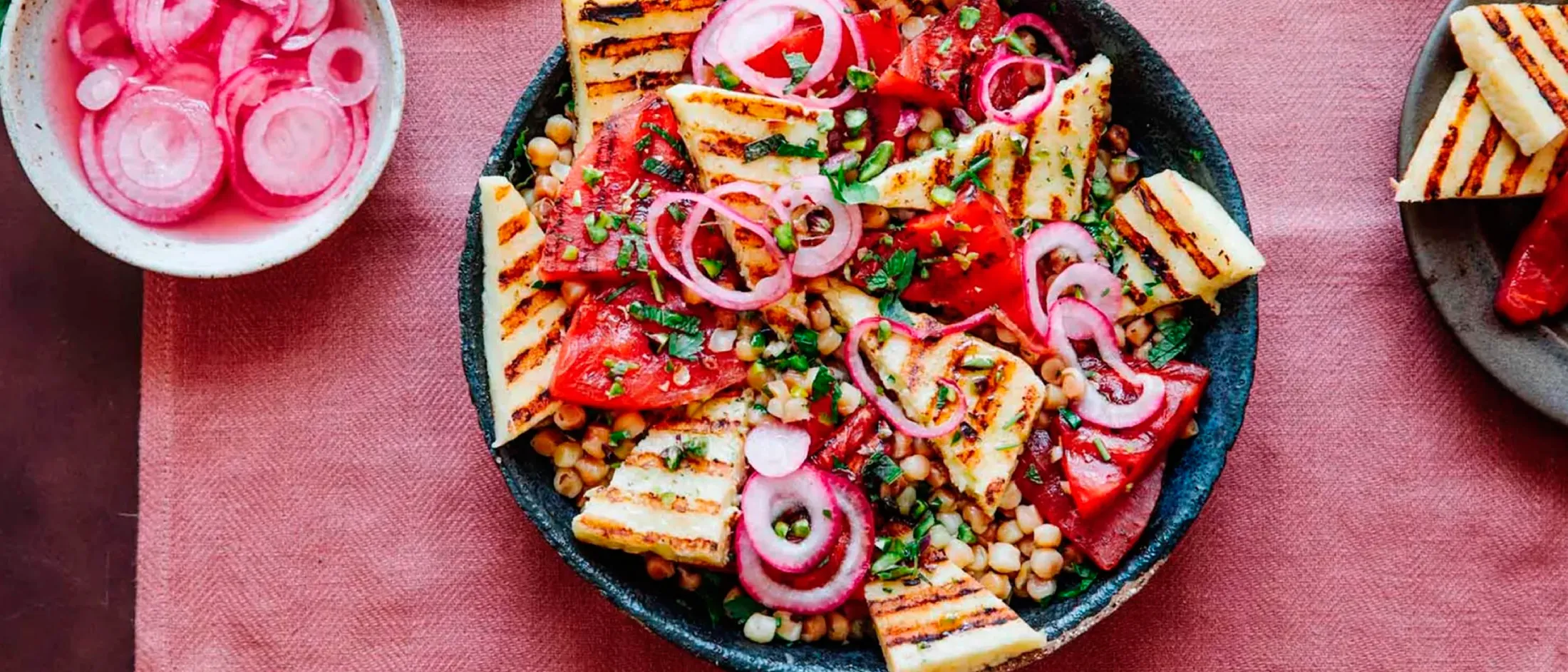 A vibrant salad featuring grilled halloumi cheese, fresh tomatoes, chickpeas, and pickled red onions, garnished with herbs.