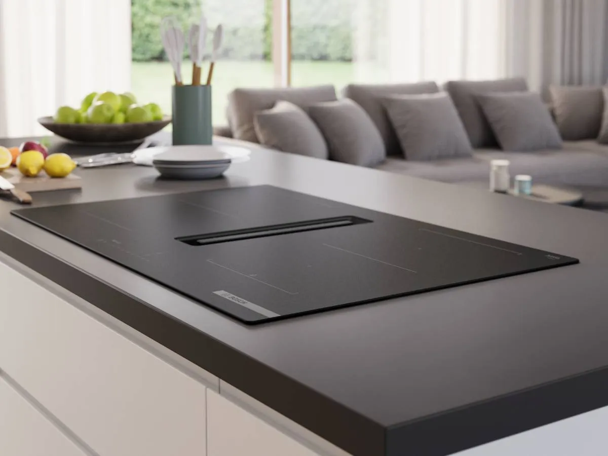 A black Bosch induction cooktop with an integrated downdraft extractor is set into a dark grey kitchen island, with a living room visible in the background.
