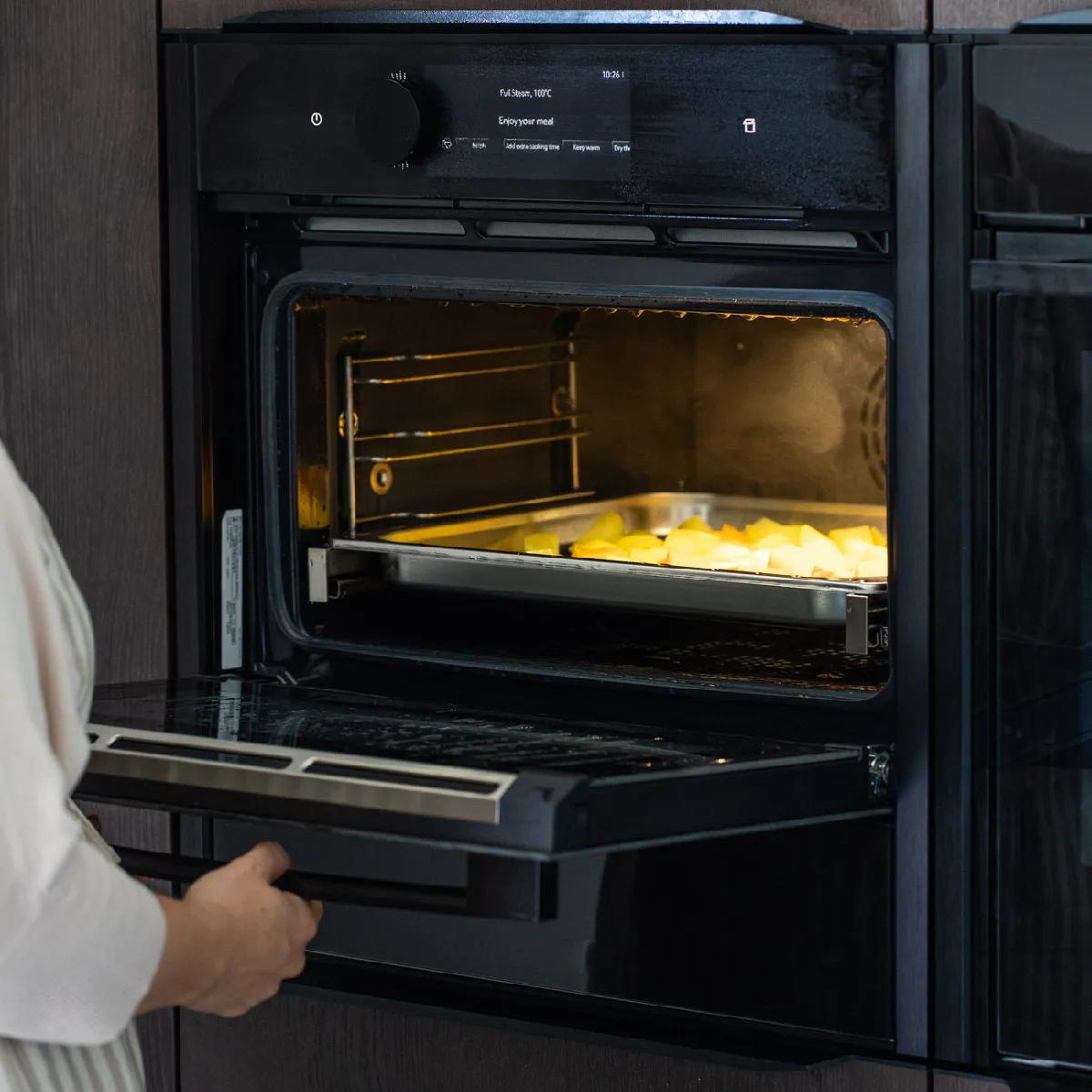 Eine Person öffnet einen modernen schwarzen Backofen, in dem ein Blech mit Kartoffelscheiben im Ofen brät; Dampf ist zu sehen.