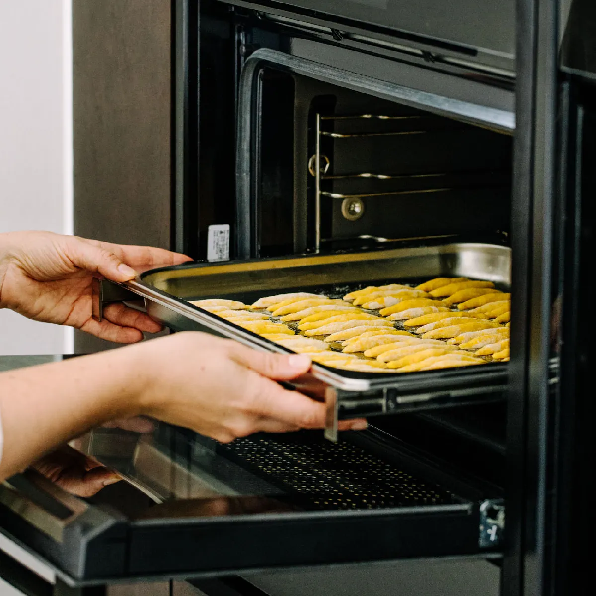 Hände, die ein Blech mit rohem Gebäck zum Backen in einen offenen Ofen schieben.